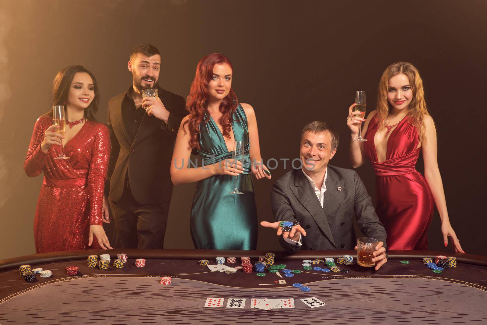 Happy colleagues are playing poker at casino. They are celebrating their win, smiling and looking vey excited while posing at the table against a dark smoke background in a ray of a spotlight. Cards, chips, money, alcohol, gambling, entertainment concept.