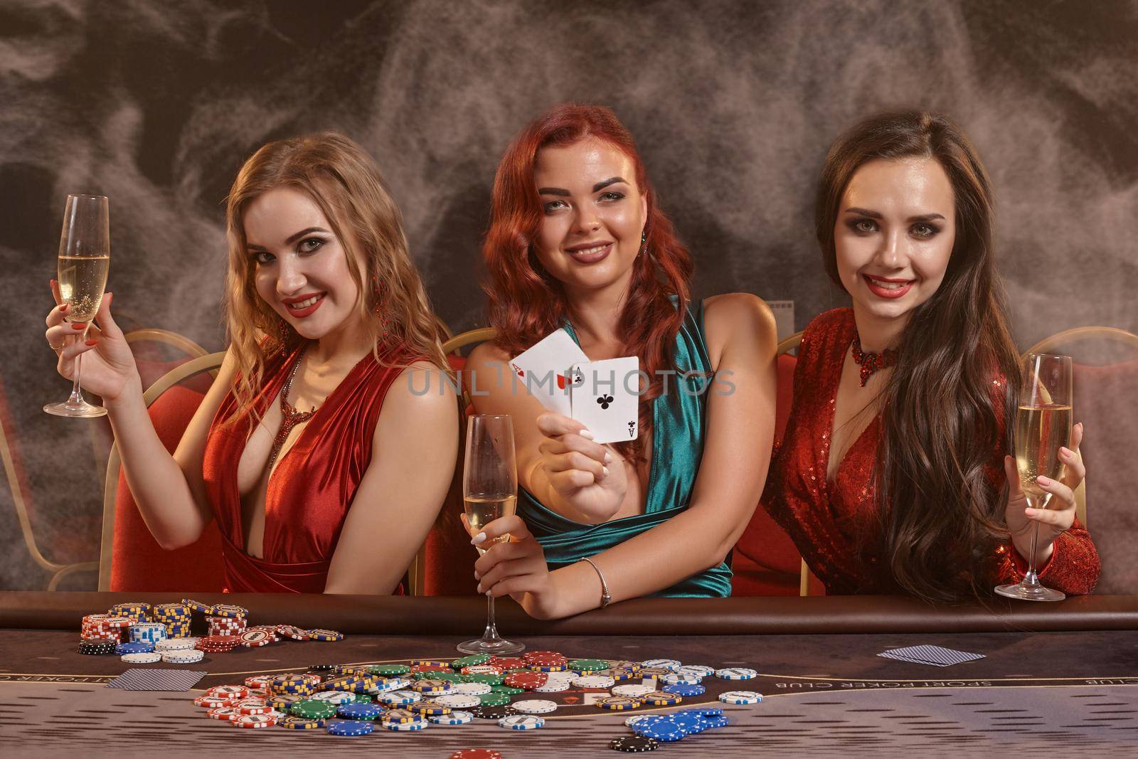 Cute maidens are playing poker at casino. They are celebrating their win, smiling, looking at the camera and posing at the table against a a dark smoke background. Cards, chips, money, alcohol, gambling, entertainment concept.