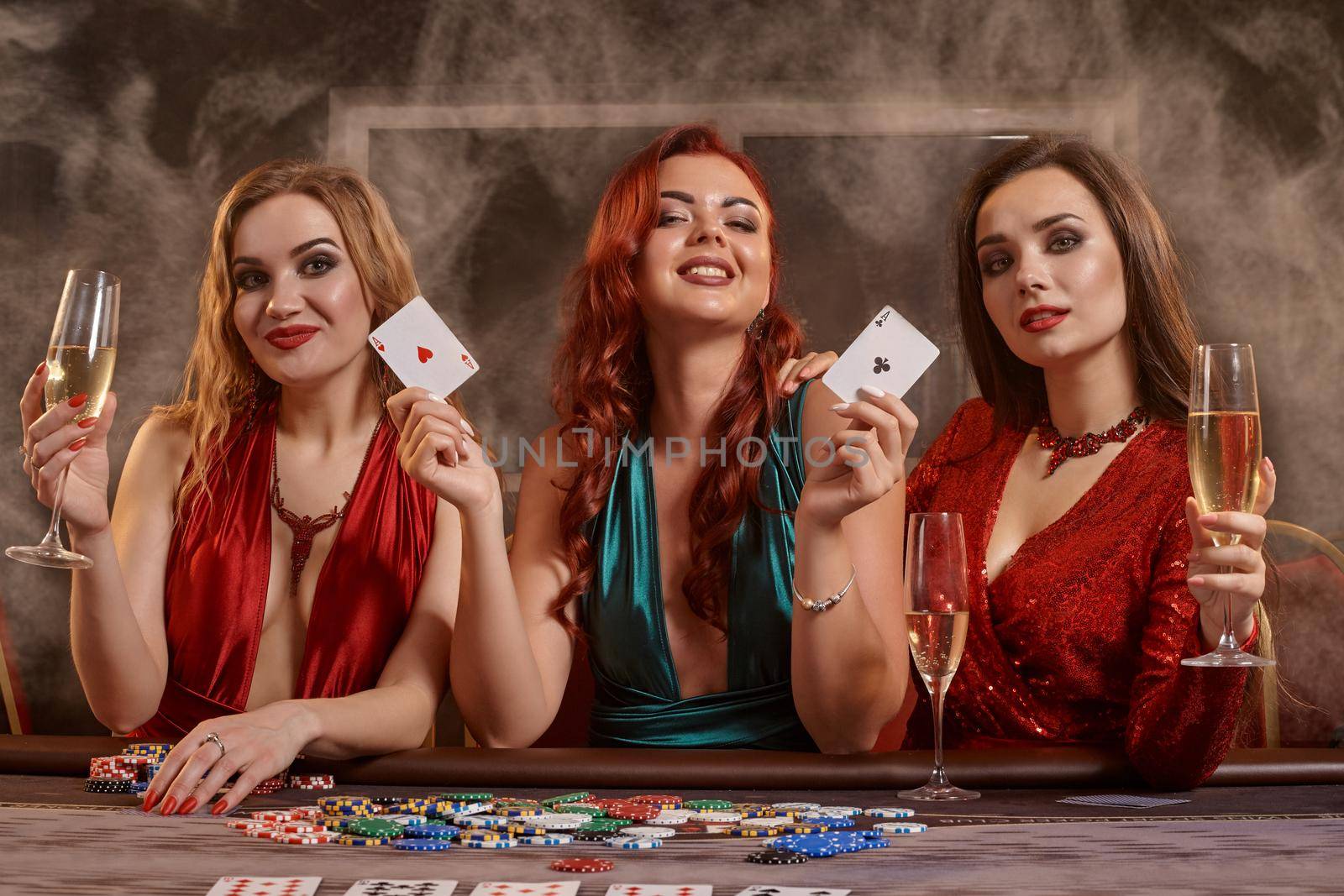 Pretty ladies are playing poker at casino. They are celebrating their win, smiling, looking at the camera and posing at the table against a a dark smoke background. Cards, chips, money, alcohol, gambling, entertainment concept.