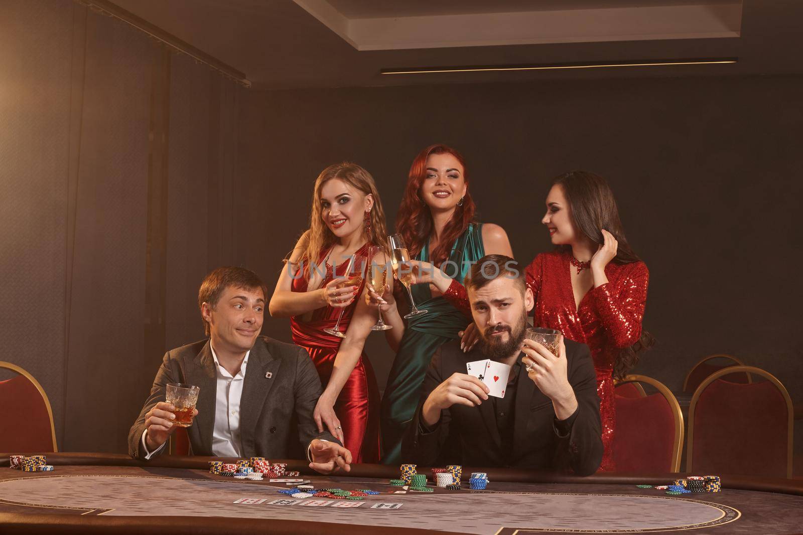 Cheerful buddies are playing poker at casino. They are celebrating their win, smiling and posing at the table against a dark smoke background. Cards, chips, money, alcohol, fortune, gambling, entertainment concept.