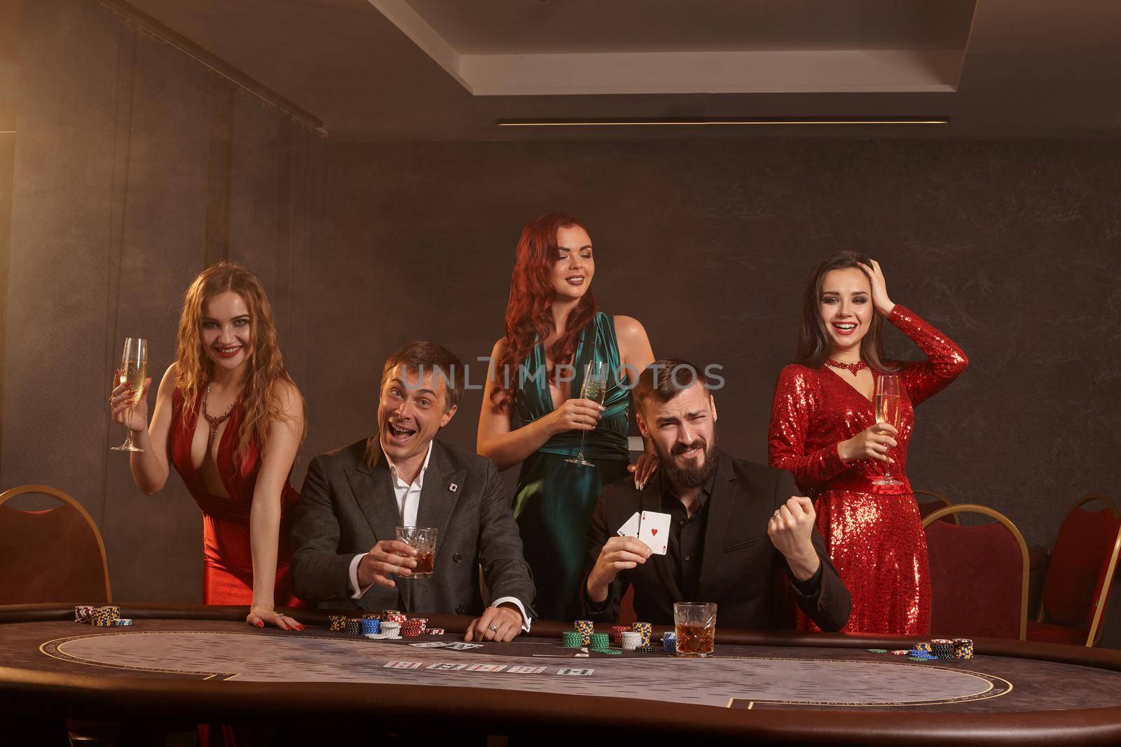 Cheerful friends are playing poker at casino. They are celebrating their win, smiling and posing at the table against a dark smoke background. Cards, chips, money, alcohol, fortune, gambling, entertainment concept.