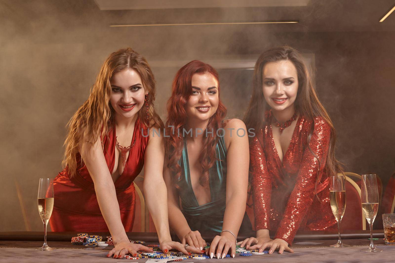 Three beautiful girls in a sexy dresses are playing poker at casino. They are celebrating their win, looking at the camera and smiling, holding some chips and posing at the table against a smoke background. Cards, money, alcohol, gambling, entertainment concept.