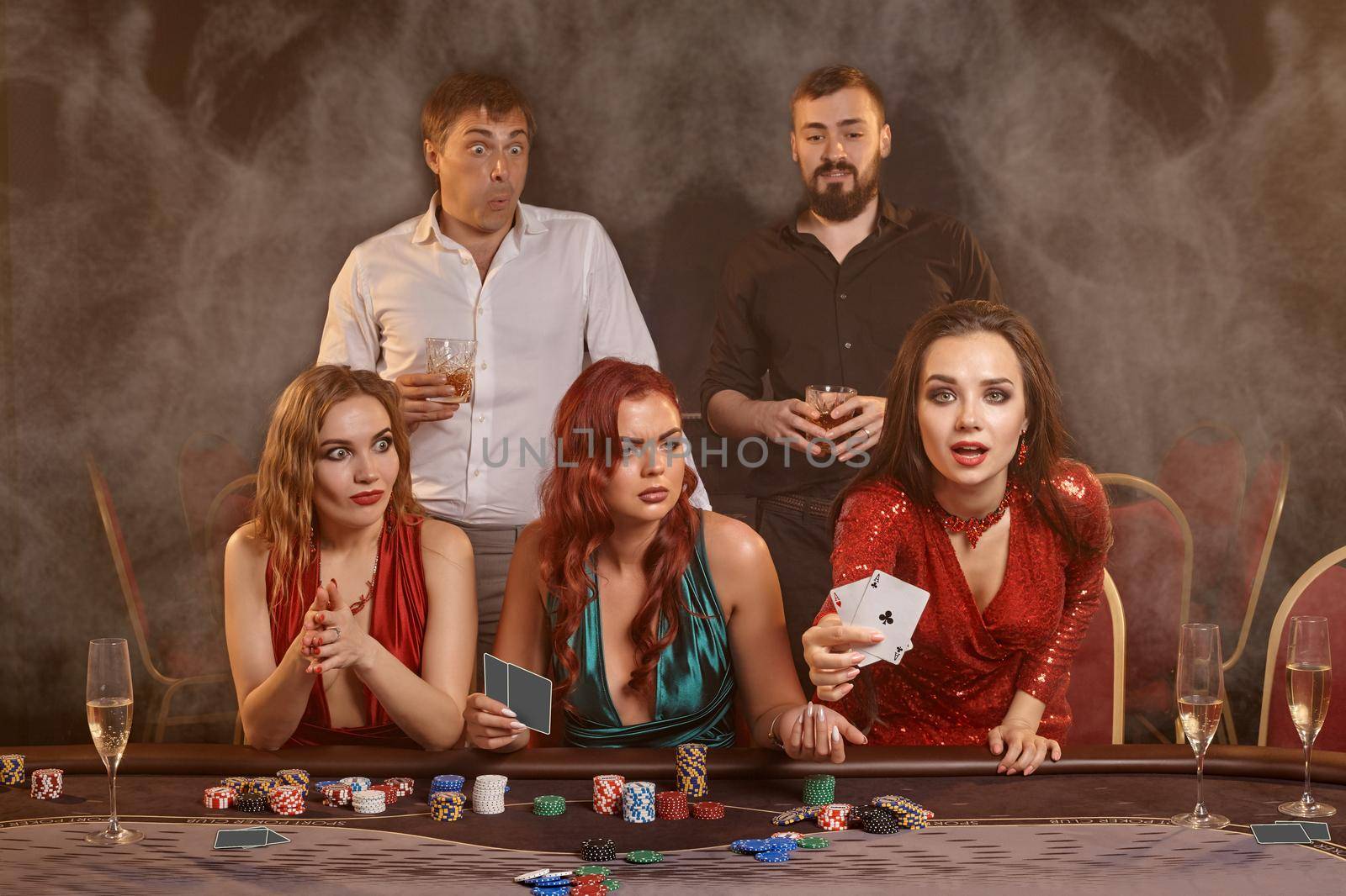 Agitated buddies are playing poker at casino. They are celebrating their win, smiling and posing at the table against a dark smoke background. Cards, chips, money, alcohol, fortune, gambling, entertainment concept.