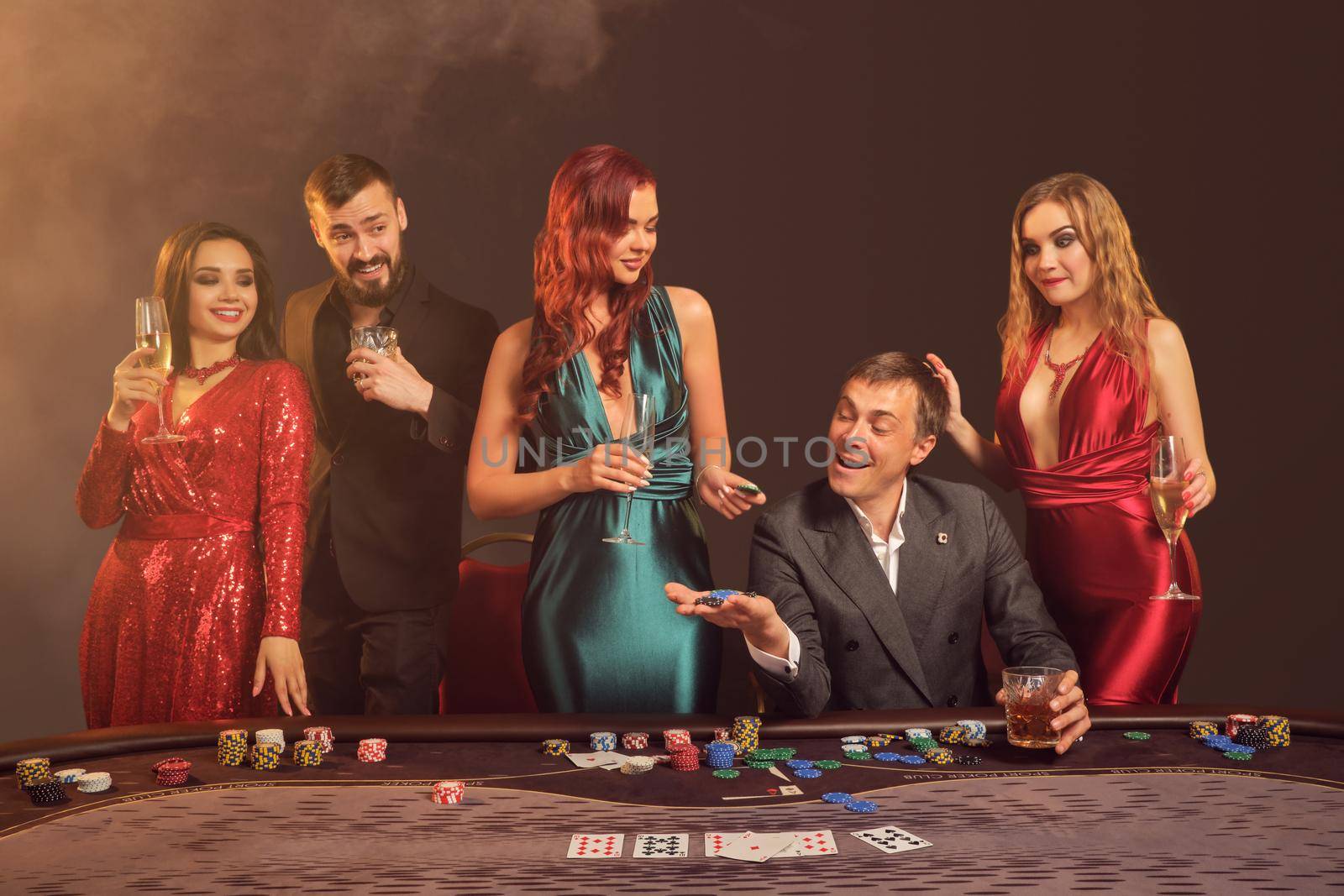 Happy classmates are playing poker at casino. They are celebrating their win, smiling and looking vey excited while posing at the table against a dark smoke background in a ray of a spotlight. Cards, chips, money, alcohol, gambling, entertainment concept.