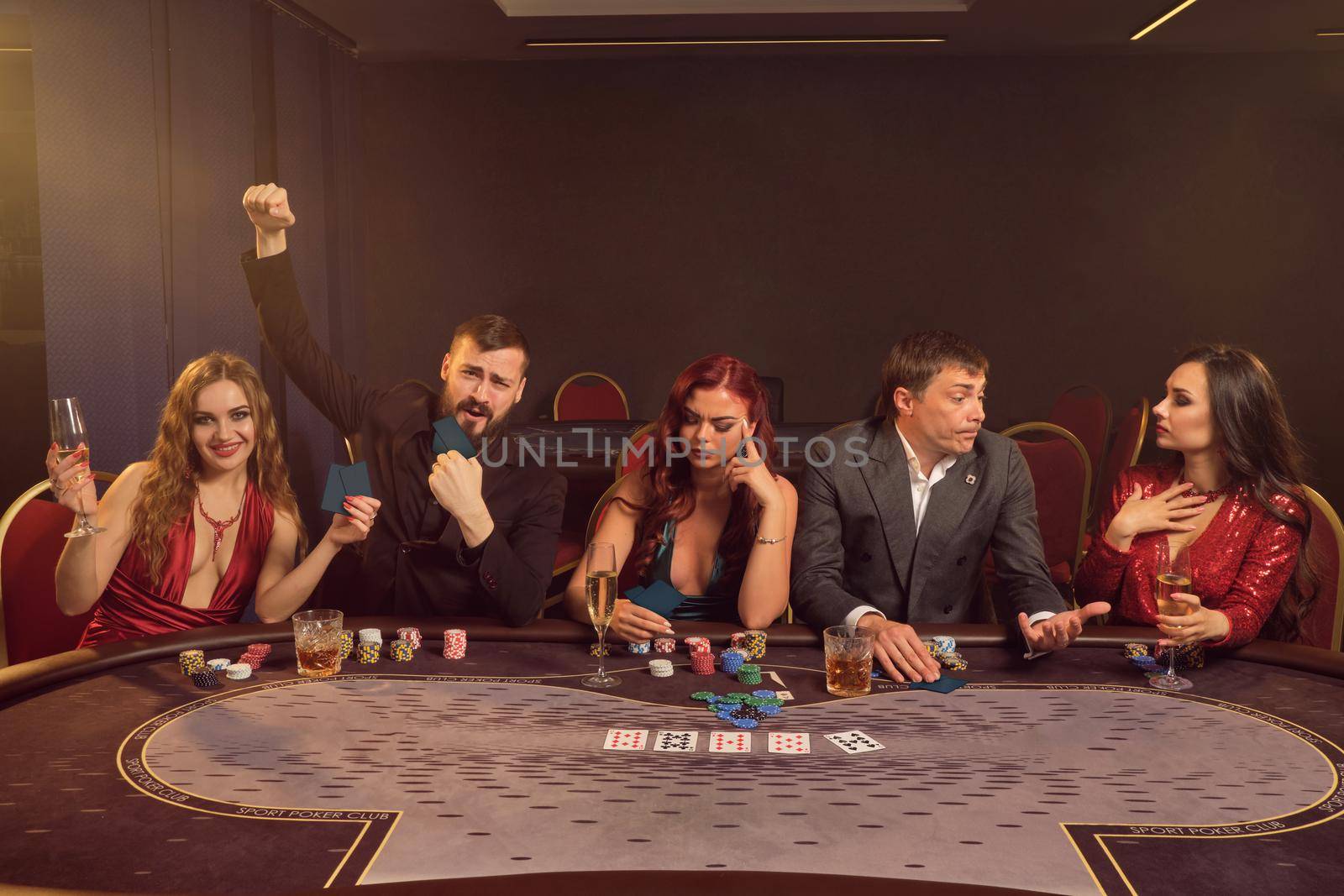 Merry classmates are playing poker at casino. They are making bets waiting for a big win while posing sitting at the table against a dark smoke background in a ray of a spotlight. Cards, chips, money, alcohol, gambling, entertainment concept.