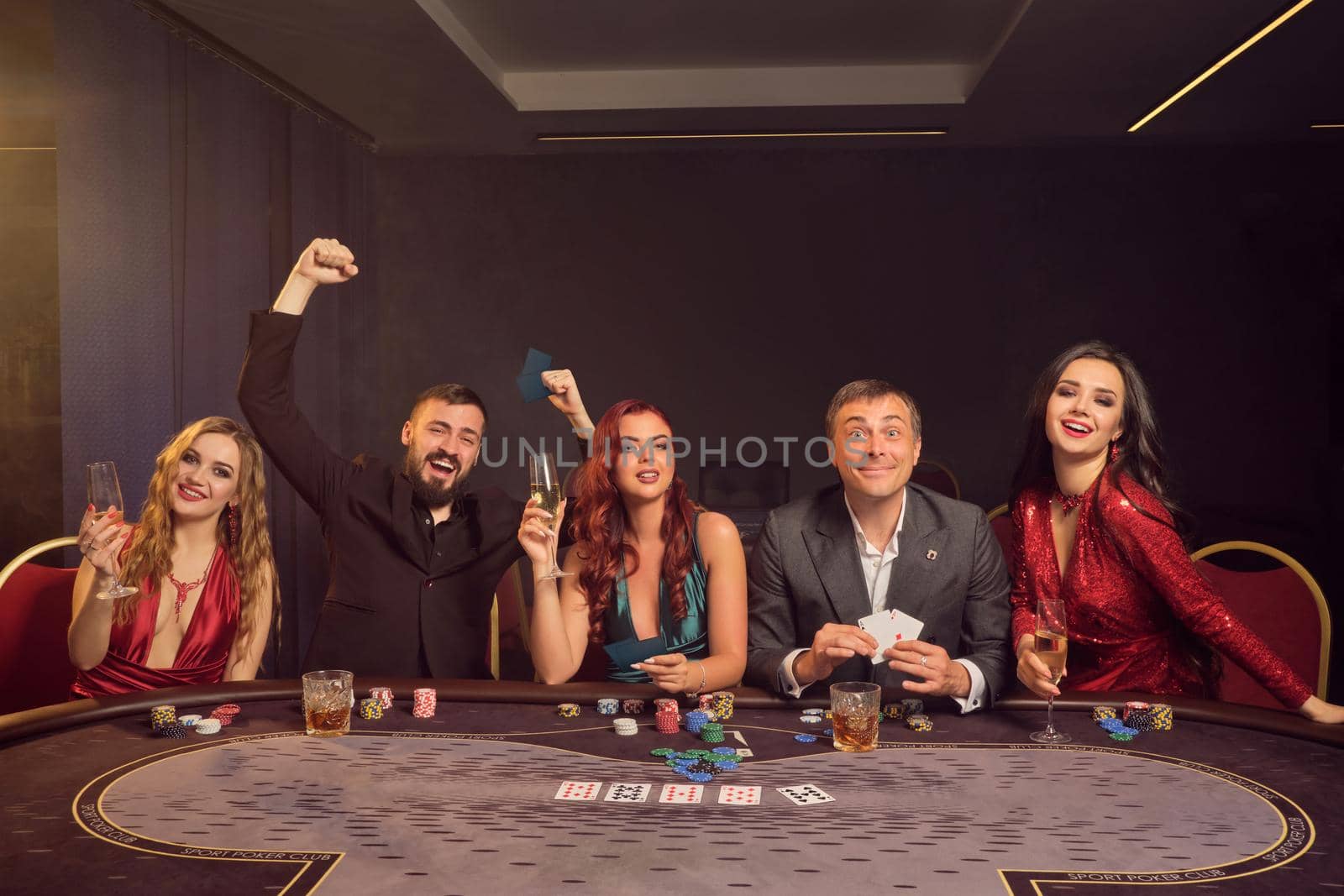 Optimistic buddies are playing poker at casino. They are celebrating their win, smiling and looking vey excited while posing sitting at the table against a dark smoke background in a ray of a spotlight. Cards, chips, money, alcohol, gambling, entertainment concept.