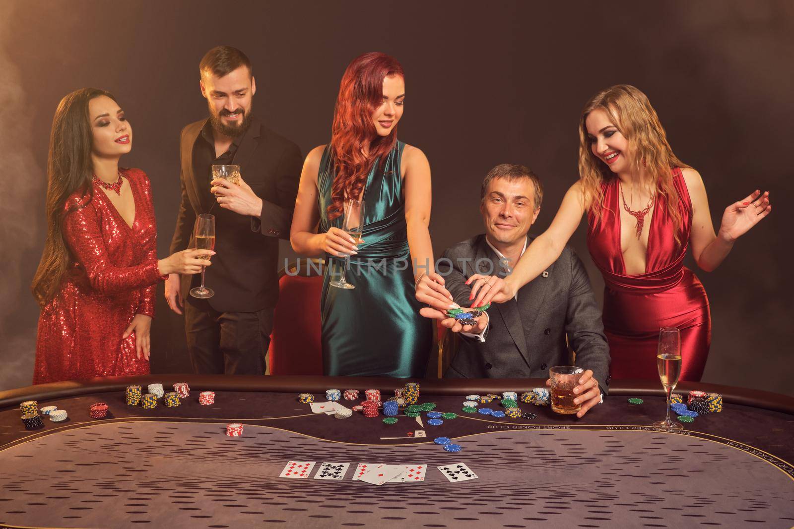 Happy buddies are playing poker at casino. They are celebrating their win, smiling and looking vey excited while posing at the table against a dark smoke background in a ray of a spotlight. Cards, chips, money, alcohol, gambling, entertainment concept.