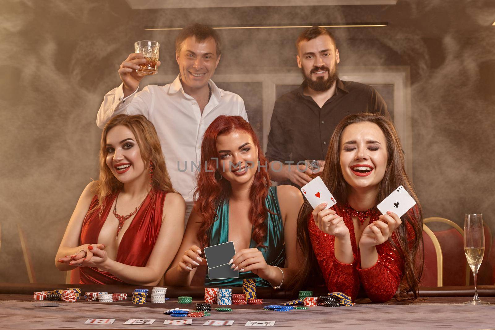Joyful classmates are playing poker at casino. They are celebrating their win, smiling and posing at the table against a dark smoke background. Cards, chips, money, alcohol, fortune, gambling, entertainment concept.