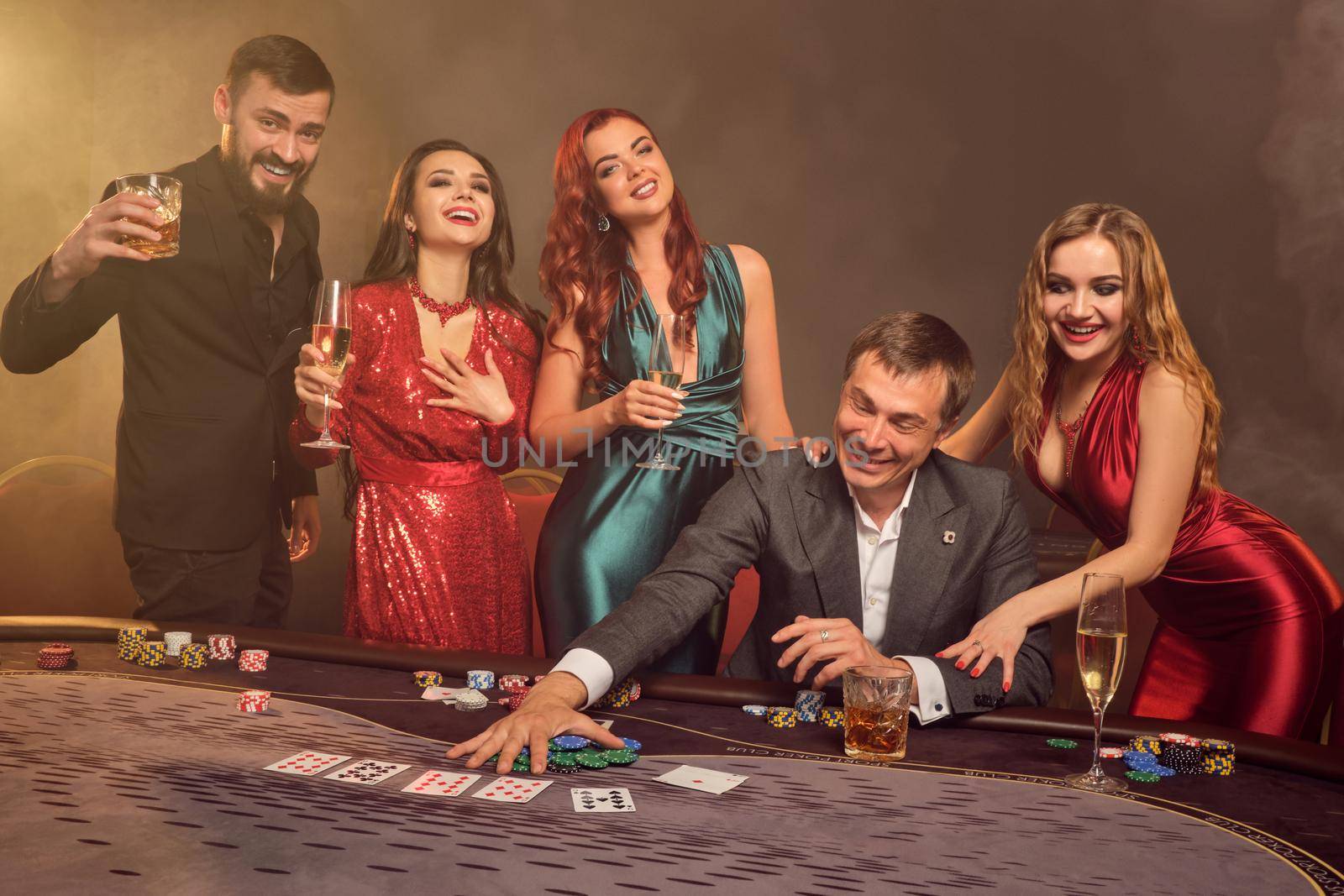Enthusiastic colleagues are playing poker at casino. They are celebrating their win, smiling and looking vey excited while posing at the table against a dark smoke background in a ray of a spotlight. Cards, chips, money, alcohol, gambling, entertainment concept.