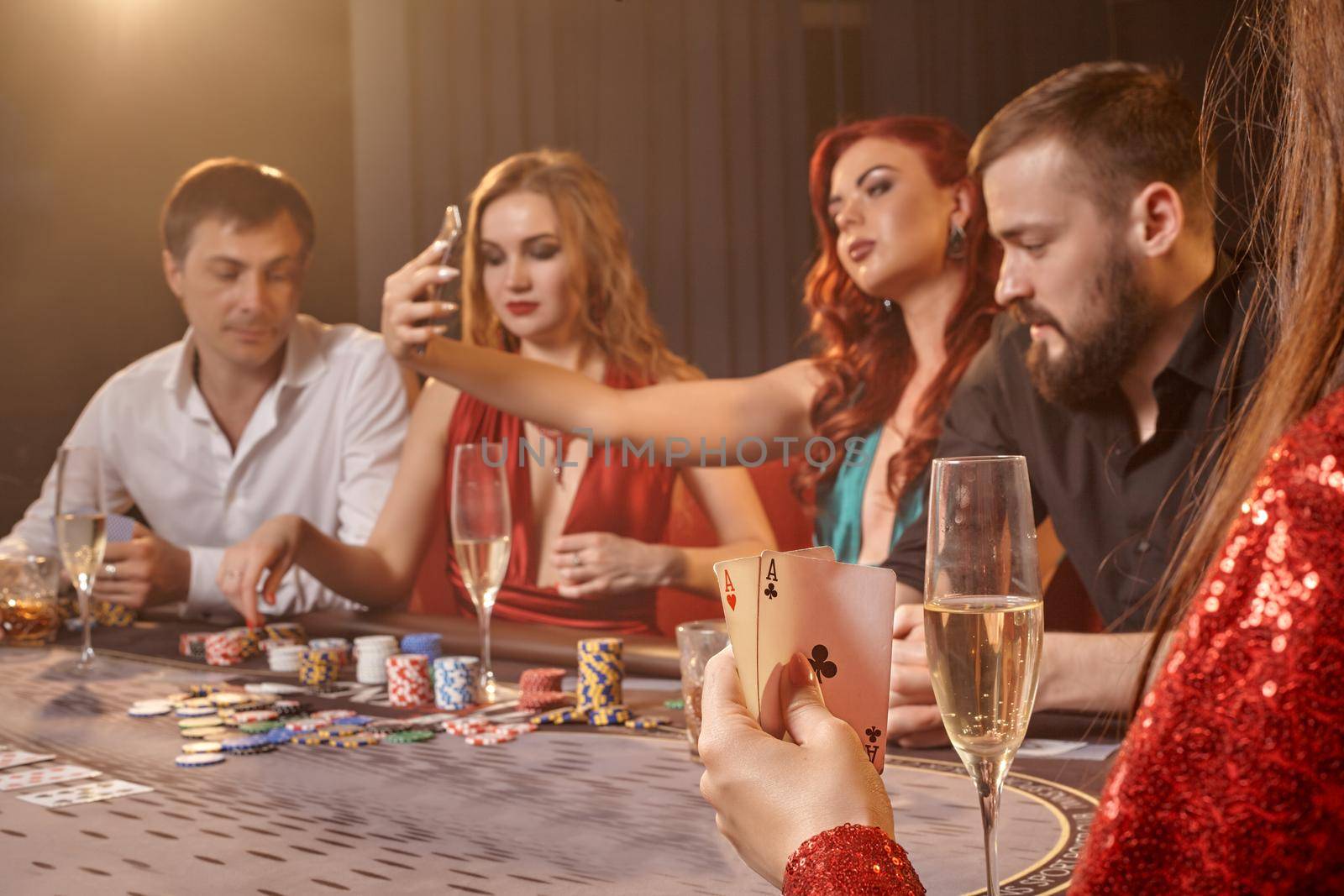 Pleasant colleagues are playing poker at casino. Youth are making bets waiting for a big win while posing at the table against a white spotlight on a dark smoke background. Cards, chips, money, alcohol, gambling, entertainment concept.