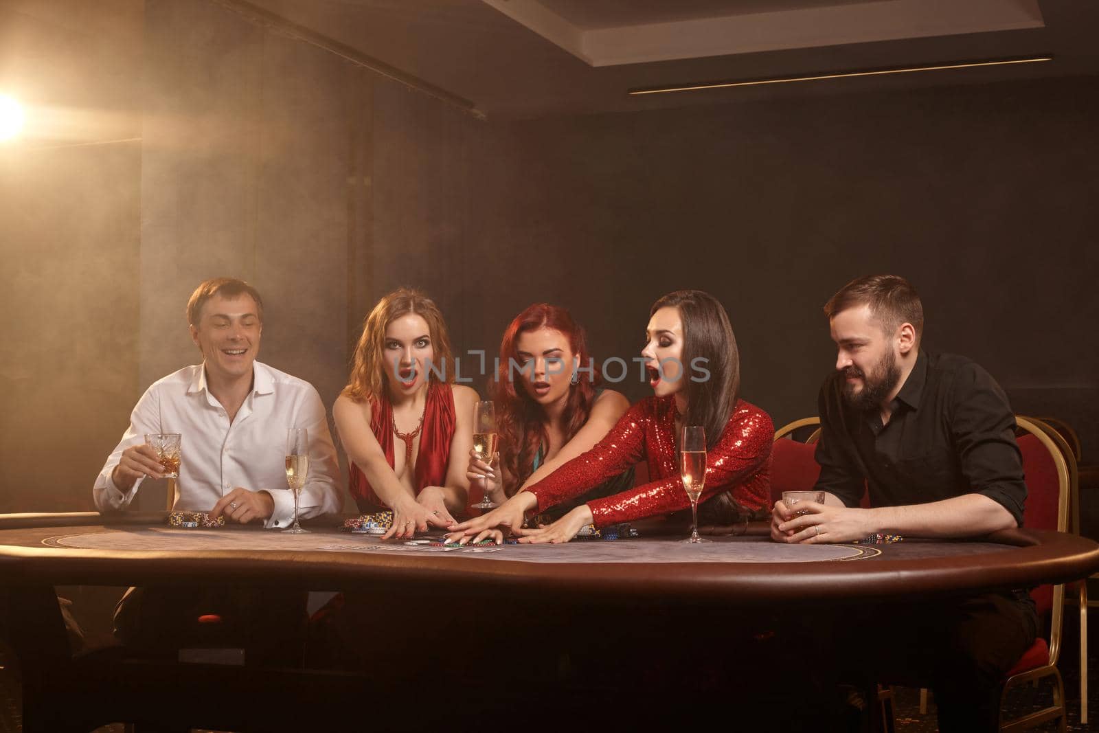 Excited friends are playing poker at casino. Golden youth are making bets waiting for a big win while posing at the table against a white spotlight on a dark smoke background. Cards, chips, money, fortune, alcohol, gambling, entertainment concept.