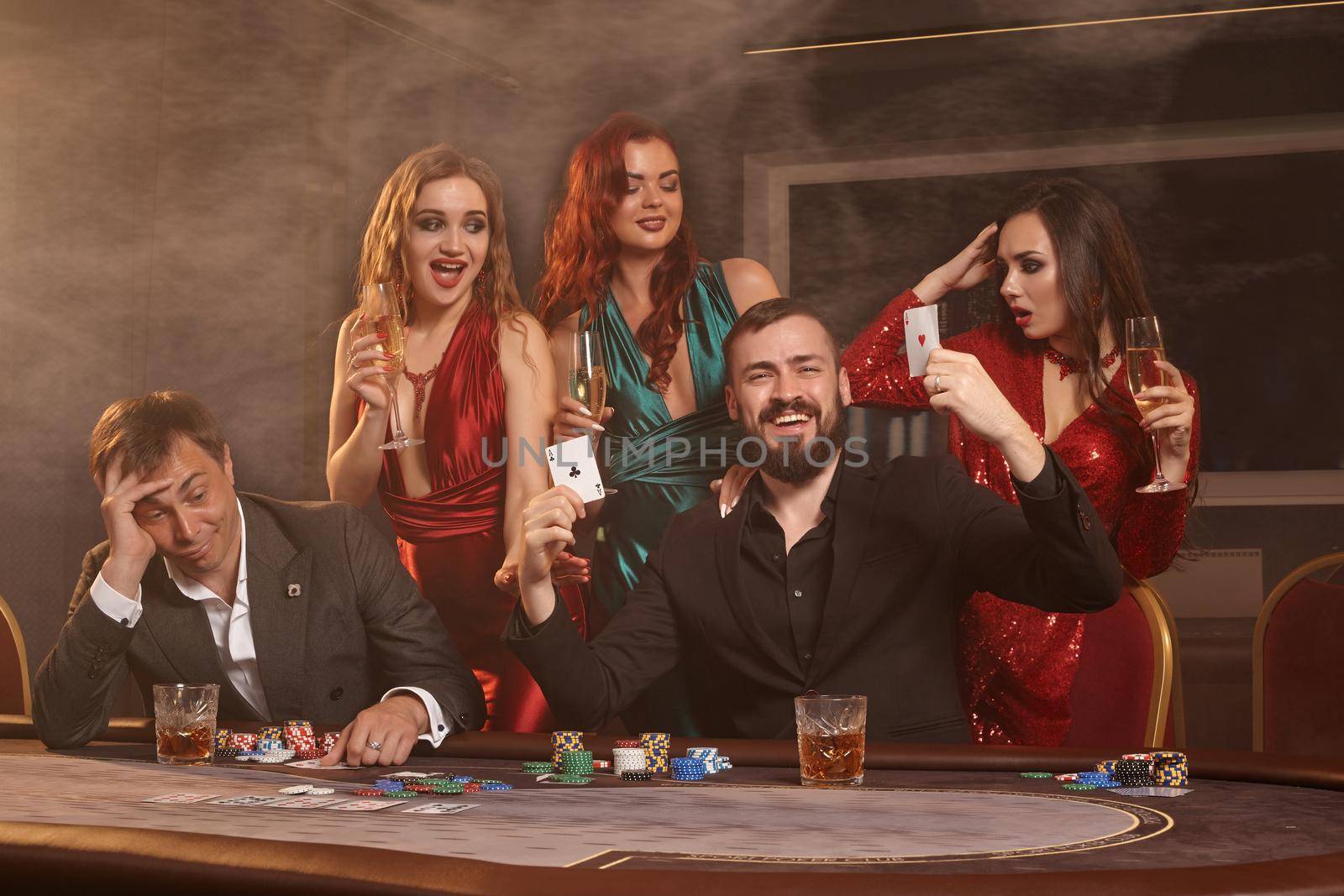 Excited colleagues are playing poker at casino. They are celebrating their win, smiling and posing at the table against a dark smoke background. Cards, chips, money, alcohol, fortune, gambling, entertainment concept.