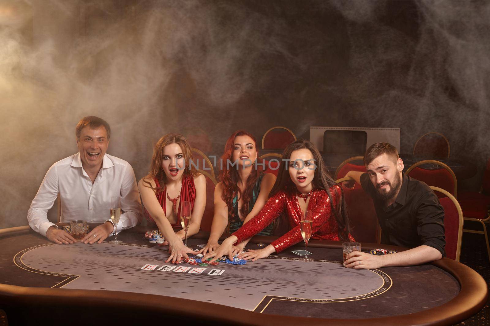 Cheerful colleagues are playing poker at casino. They are celebrating their win, smiling and looking vey excited while posing at the table against a dark smoke background in a ray of a spotlight. Cards, chips, money, alcohol, gambling, entertainment concept.