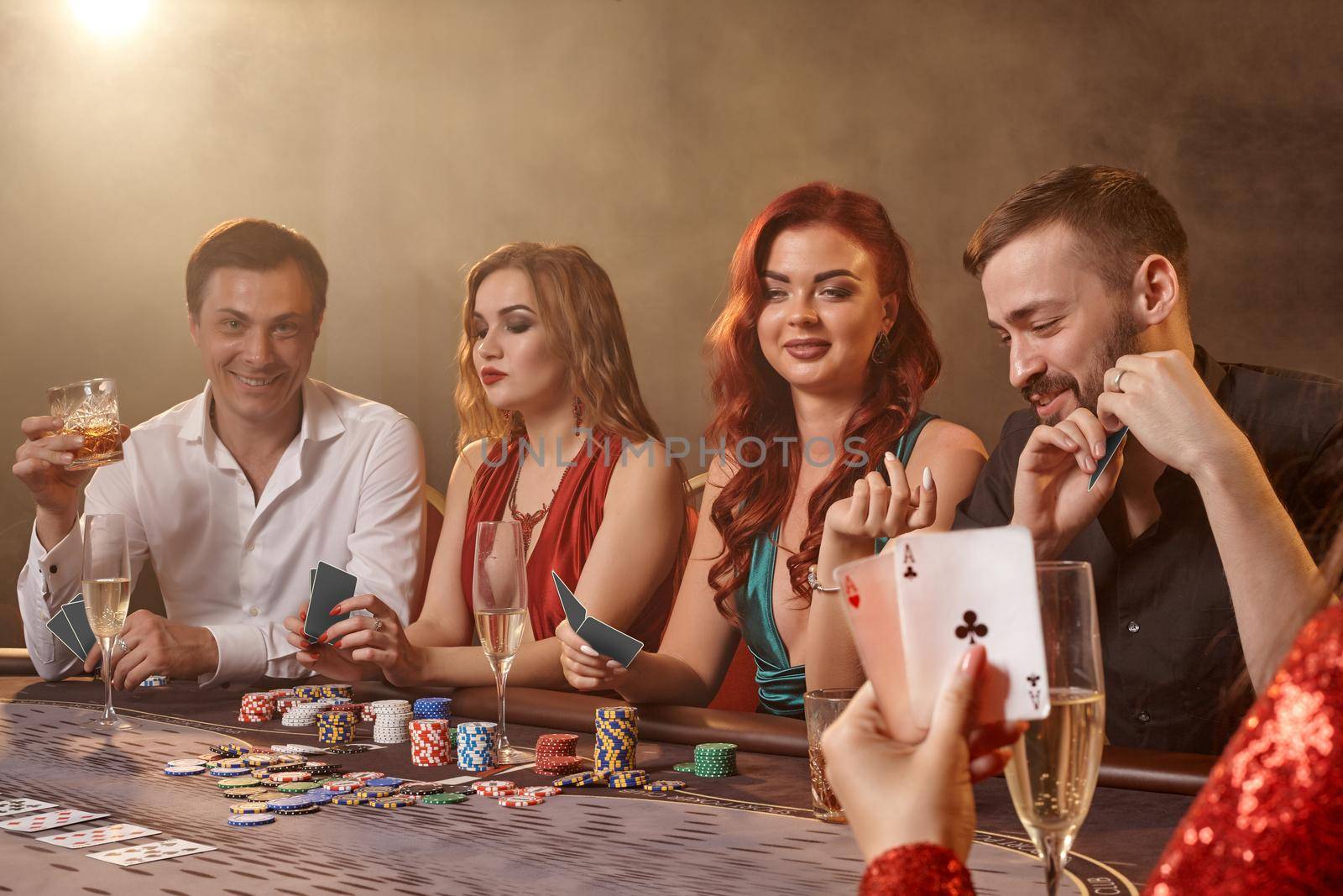 Pleasant friends are playing poker at casino. Youth are making bets waiting for a big win while posing at the table against a white spotlight on a dark smoke background. Cards, chips, money, alcohol, gambling, entertainment concept.