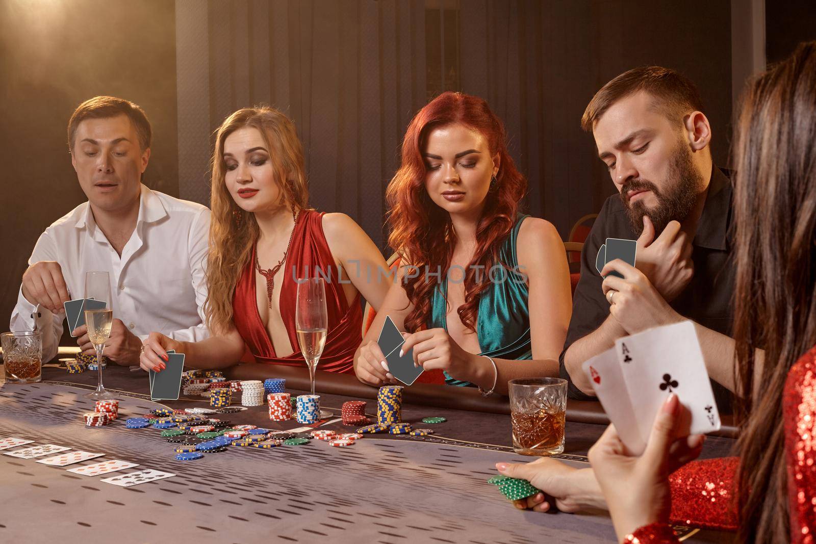 Enthusiastic classmates are playing poker at casino. Youth are making bets waiting for a big win while posing at the table against a white spotlight on a dark smoke background. Cards, chips, money, alcohol, gambling, entertainment concept.