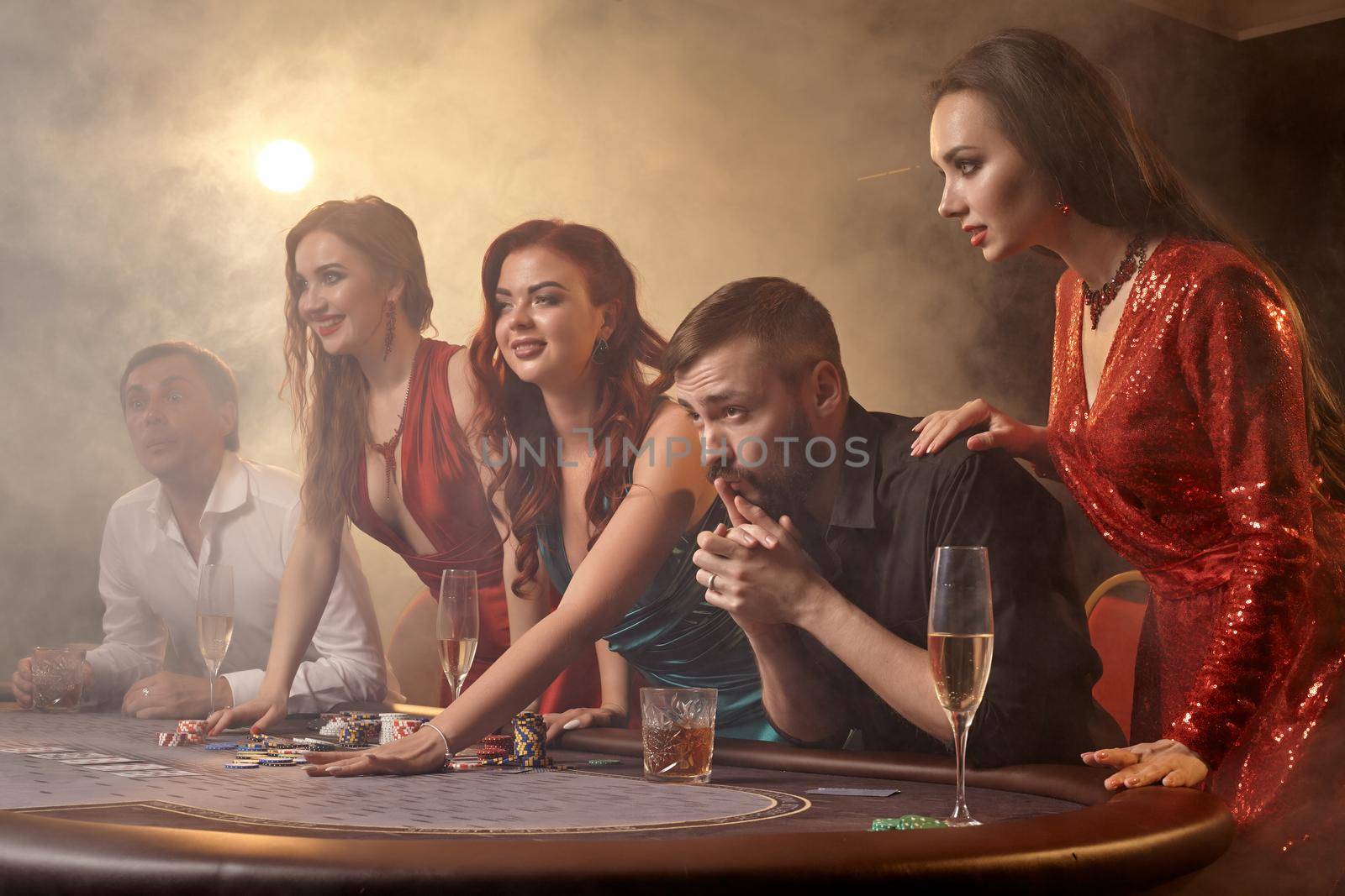 Cheerful companions are playing poker at casino. Youth are making bets waiting for a big win while posing at the table against a white spotlight on a dark smoke background. Cards, chips, money, alcohol, gambling, entertainment concept.
