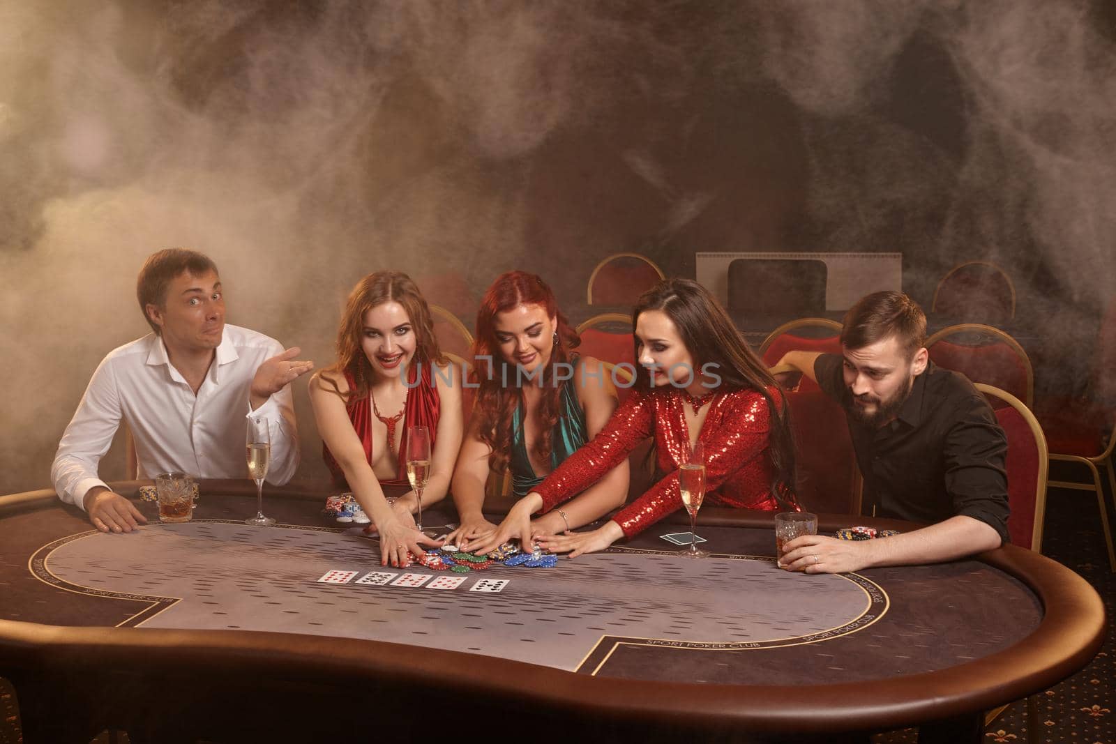 Wealthy friends are playing poker at casino. They are celebrating their win, smiling and looking vey excited while posing at the table against a dark smoke background. Cards, chips, money, alcohol, gambling, entertainment concept.