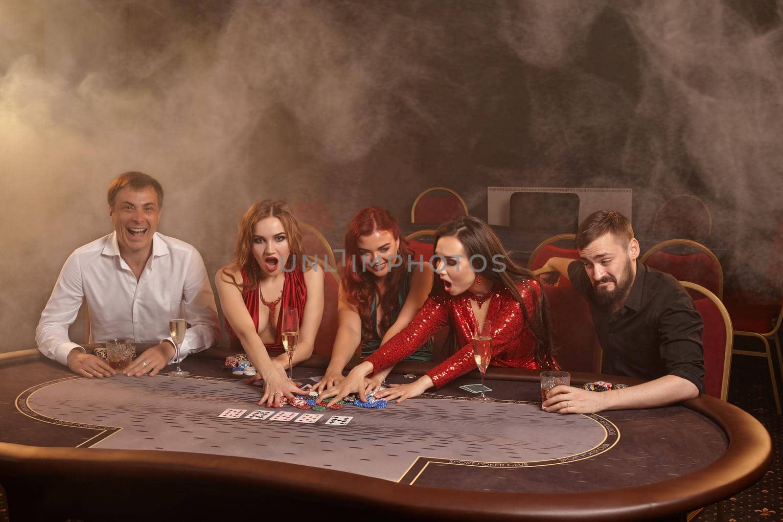 Cheerful partners are playing poker at casino. They are celebrating their win, smiling and looking vey excited while posing sitting at the table against a dark smoke background in a ray of a spotlight. Cards, chips, money, alcohol, gambling, entertainment concept.
