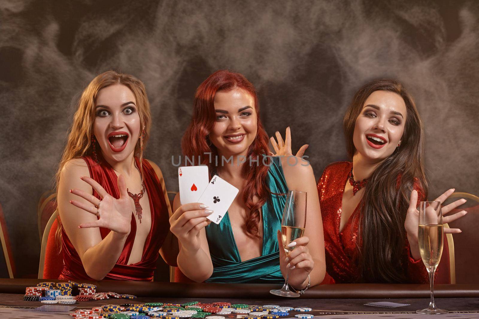 Sexy women are playing poker at casino. They are celebrating their win, smiling, looking at the camera and posing at the table against a a dark smoke background. Cards, chips, money, alcohol, gambling, entertainment concept.