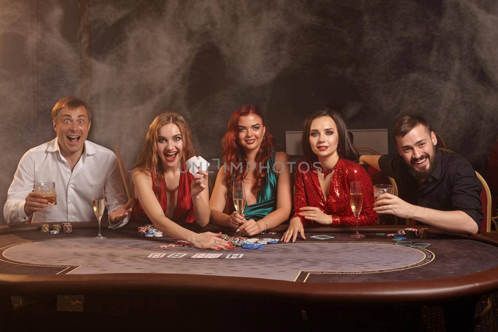 Cheerful buddies are playing poker at casino. They are celebrating their win, smiling and looking vey excited while posing at the table against a dark smoke background. Cards, chips, money, alcohol, gambling, entertainment concept.