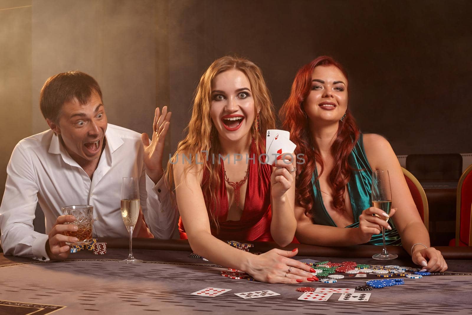 Group of a funny friends are playing poker at casino. They are celebrating their win, looking at the camera and smiling while showing the winning combination and posing sitting at the table against a dark smoke background. Cards, chips, money, gambling, entertainment concept.