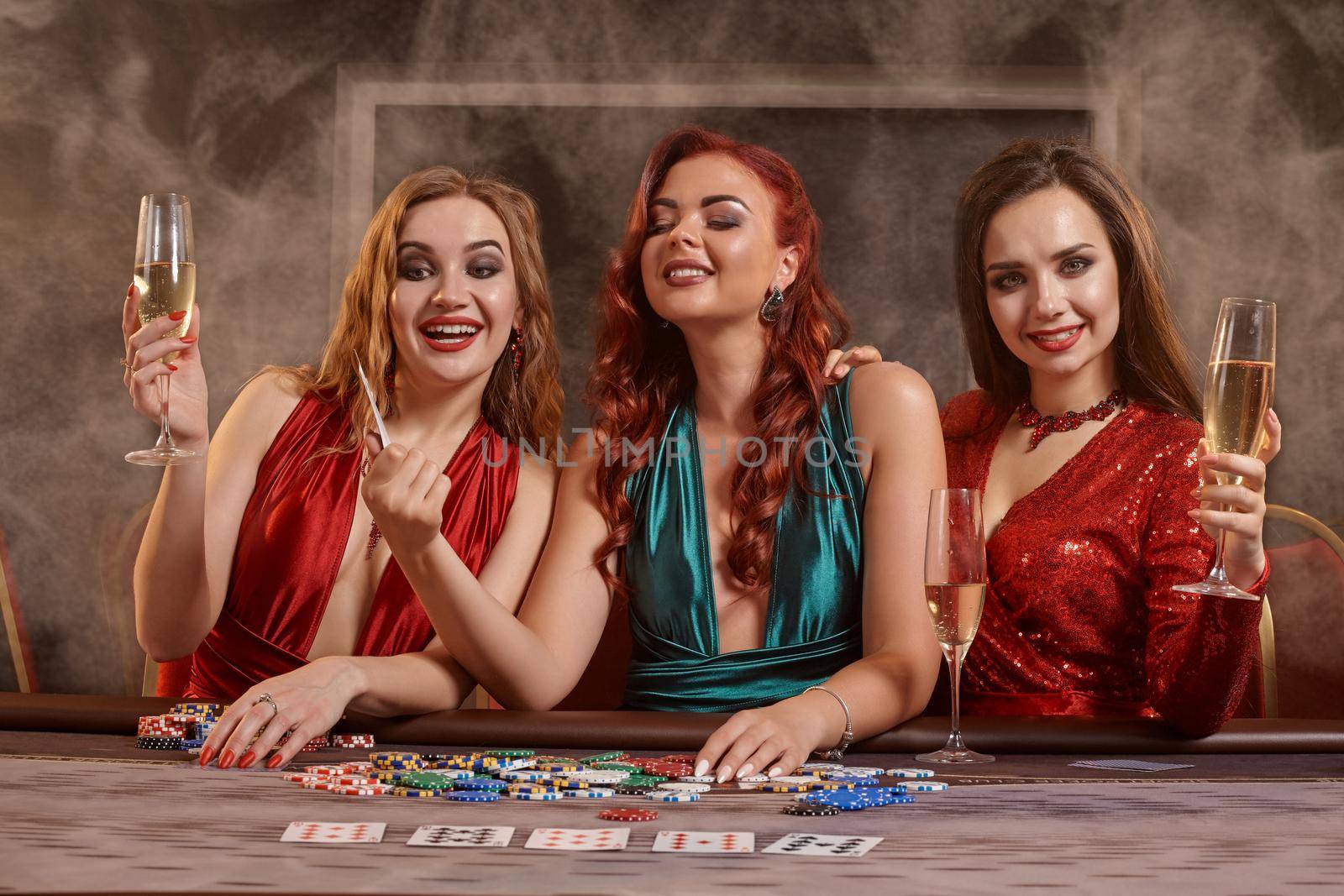 Beautiful females are playing poker at casino. They are celebrating their win, smiling, looking at the camera and posing at the table against a a dark smoke background. Cards, chips, money, alcohol, gambling, entertainment concept.