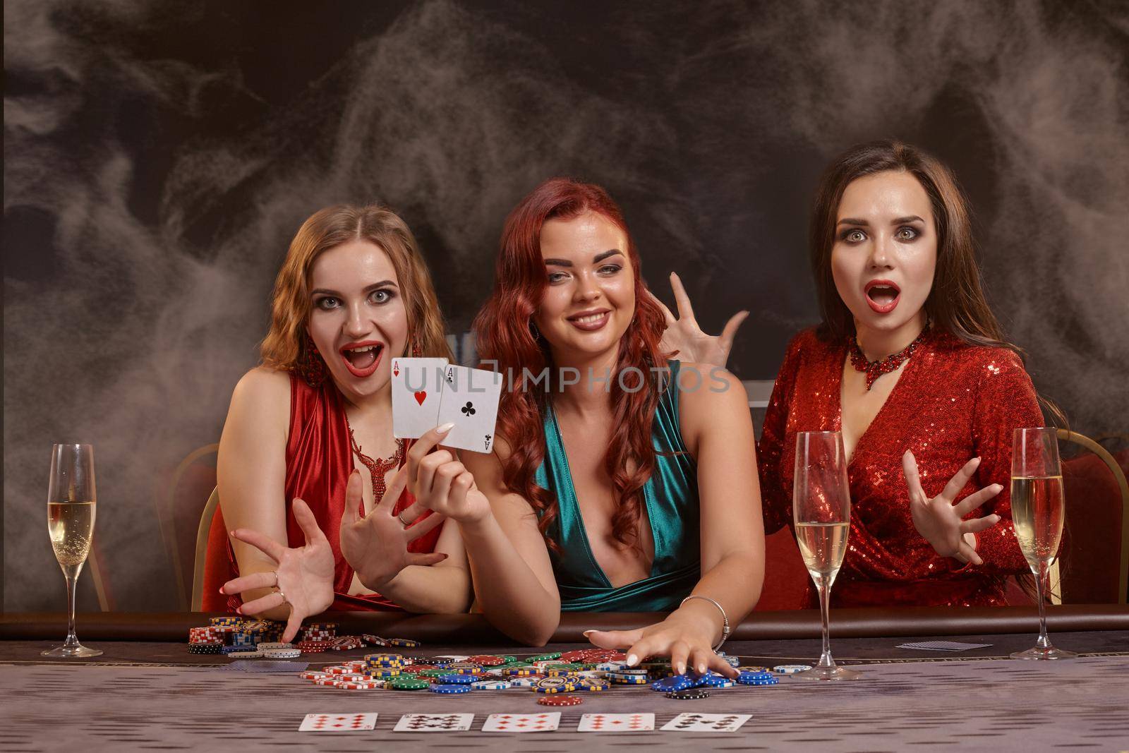 Attractive girls are playing poker at casino. They are celebrating their win, smiling, looking at the camera and posing at the table against a a dark smoke background. Cards, chips, money, alcohol, gambling, entertainment concept.