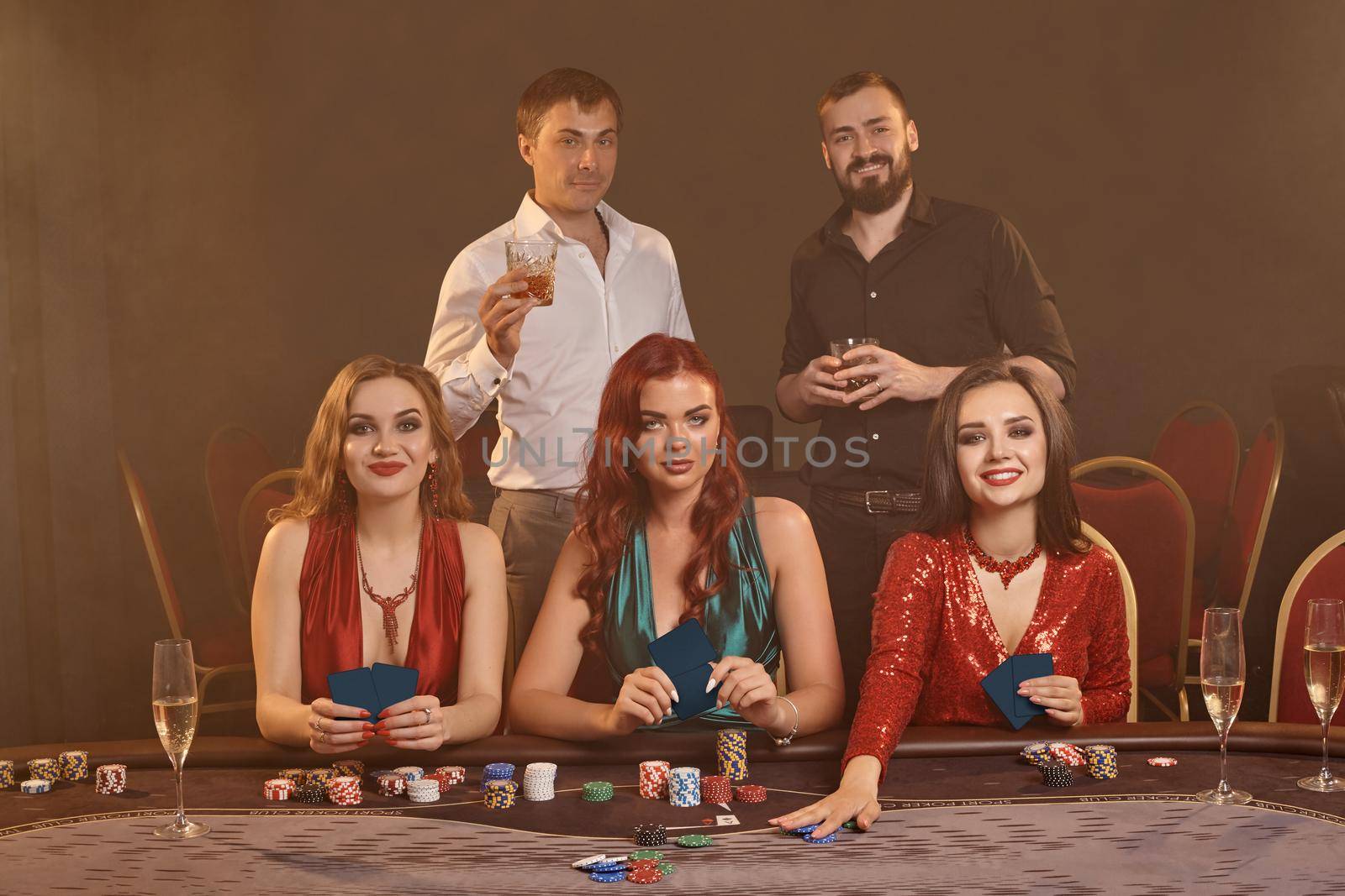 Concentrated buddies are playing poker at casino. Golden youth are making bets waiting for a big win while posing at the table against a dark smoke background. Cards, chips, money, fortune, alcohol, gambling, entertainment concept.