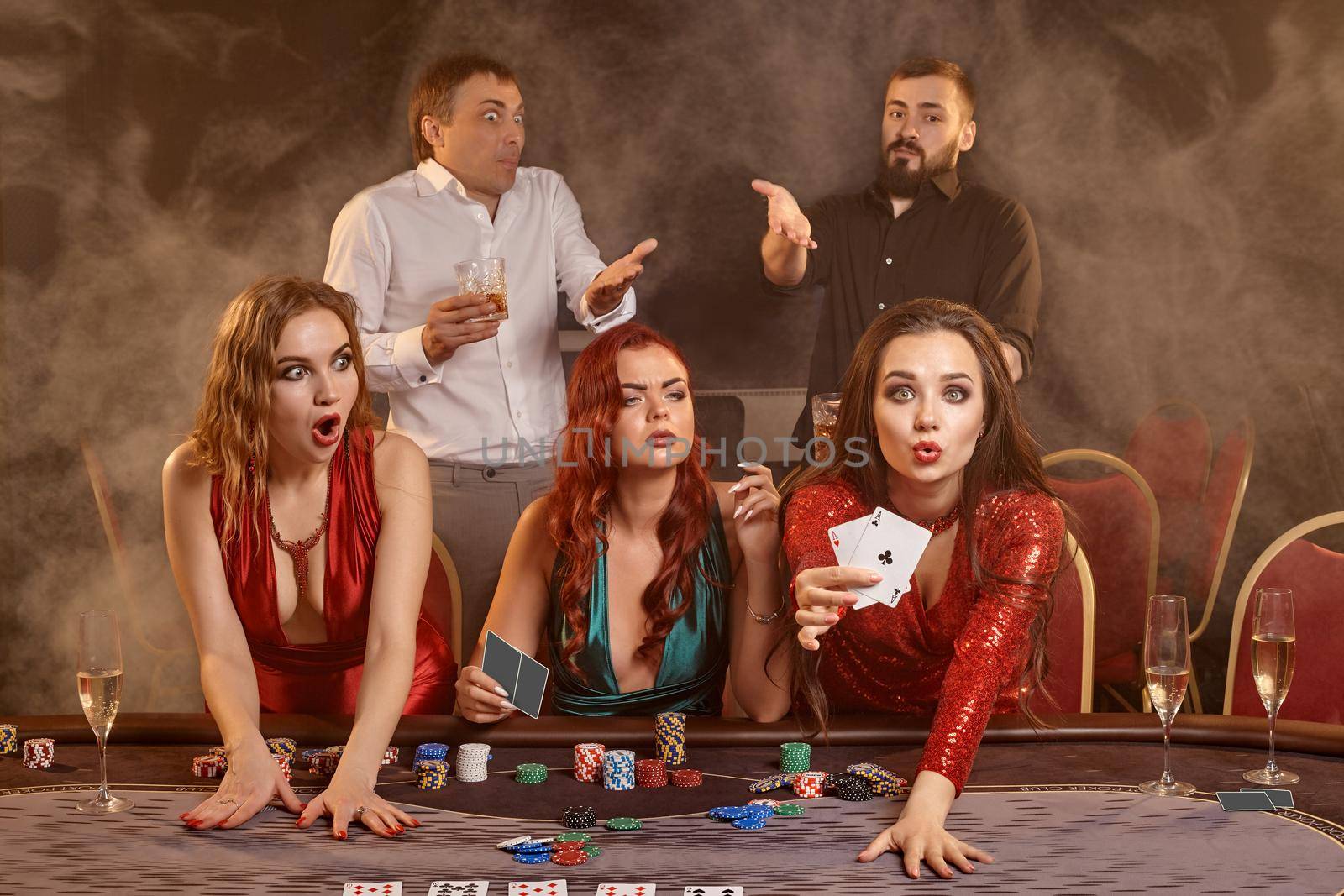 Agitated classmates are playing poker at casino. They are celebrating their win, smiling and posing at the table against a dark smoke background. Cards, chips, money, alcohol, fortune, gambling, entertainment concept.