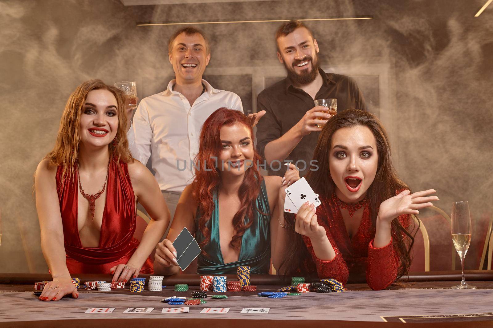 Joyful friends are playing poker at casino. They are celebrating their win, smiling and posing at the table against a dark smoke background. Cards, chips, money, alcohol, fortune, gambling, entertainment concept.