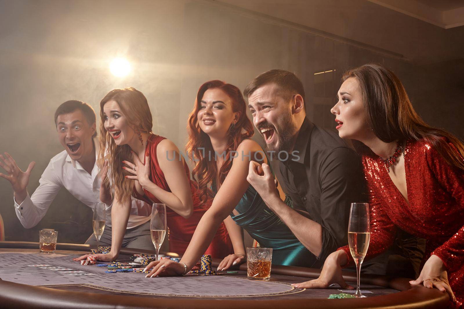 Excited friends are playing poker at casino. Youth are making bets waiting for a big win while posing at the table against a white spotlight on a dark smoke background. Cards, chips, money, alcohol, gambling, entertainment concept.