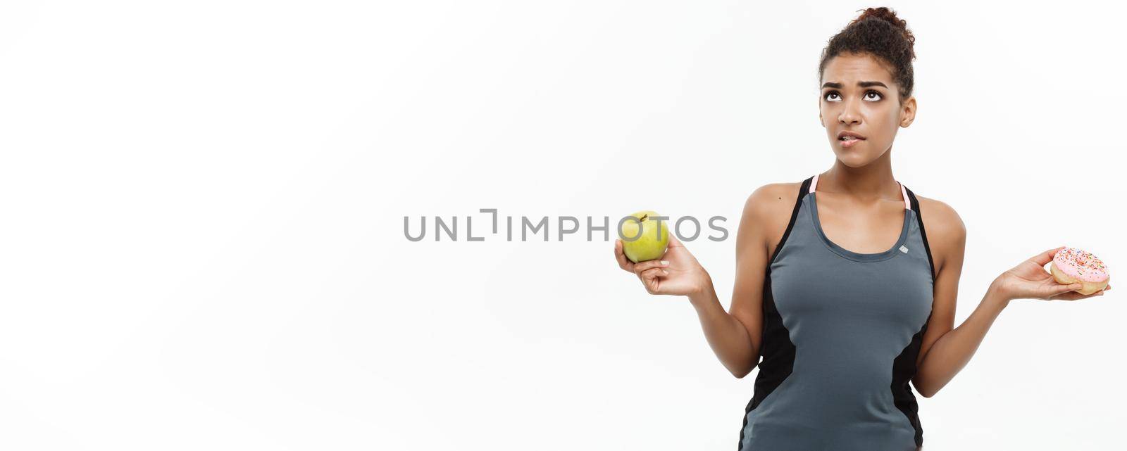 Healthy and diet concept - Beautiful sporty African American make a decision between donut and green apple. Isolated on white background. by Benzoix