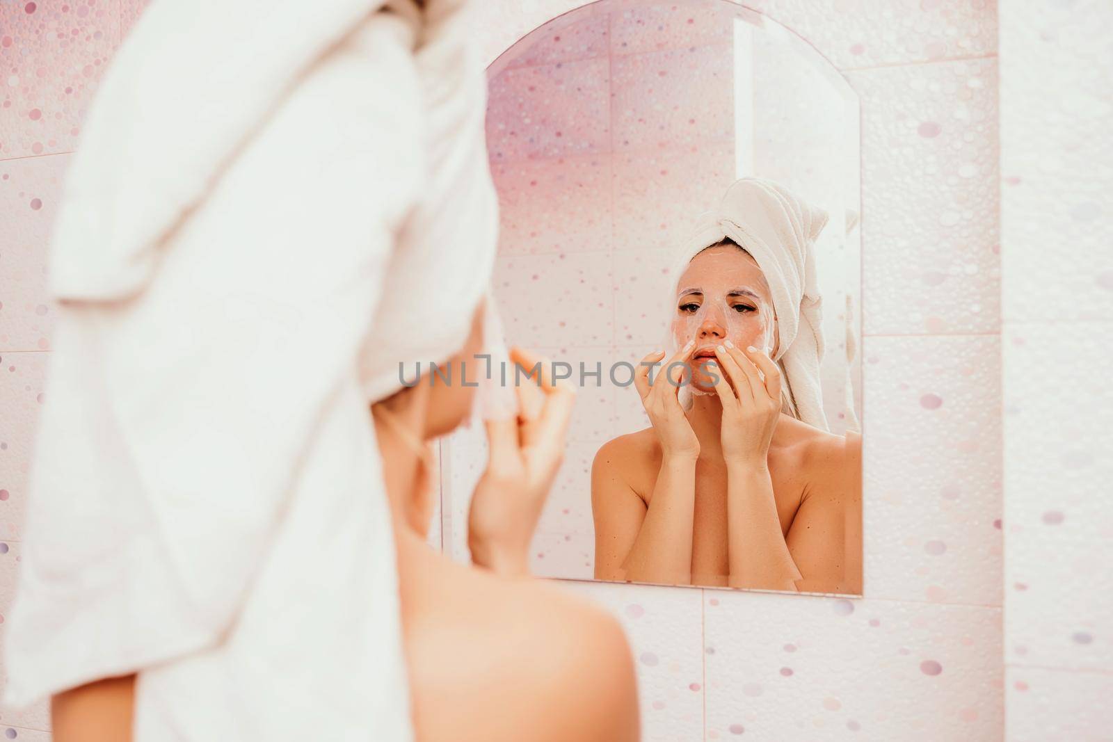 Young beautiful woman using a moisturizing facial mask after taking a bath. Pretty attractive girl in a towel on her head stands in front of a mirror in a home bathroom. Daily hygiene and skin care.