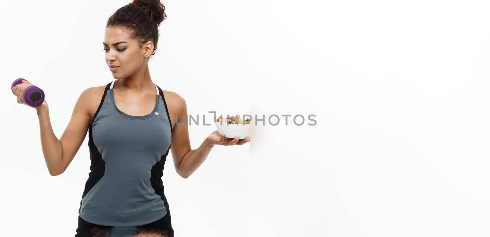 Healthy and Fitness concept - Beautiful sporty African American on diet holding dumbbell and fresh salad on hands. Isolated on white studio background