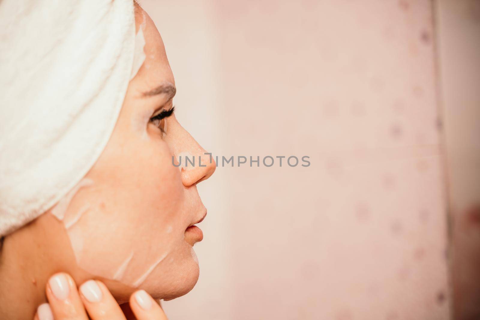 Young beautiful woman using a moisturizing facial mask after taking a bath. Pretty attractive girl in a towel on her head stands in front of a mirror in a home bathroom. Daily hygiene and skin care by Matiunina