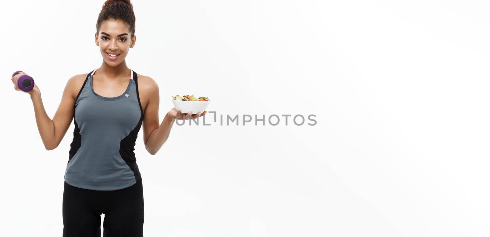 Healthy and Fitness concept - Beautiful sporty African American on diet holding dumbbell and fresh salad on hands. Isolated on white studio background