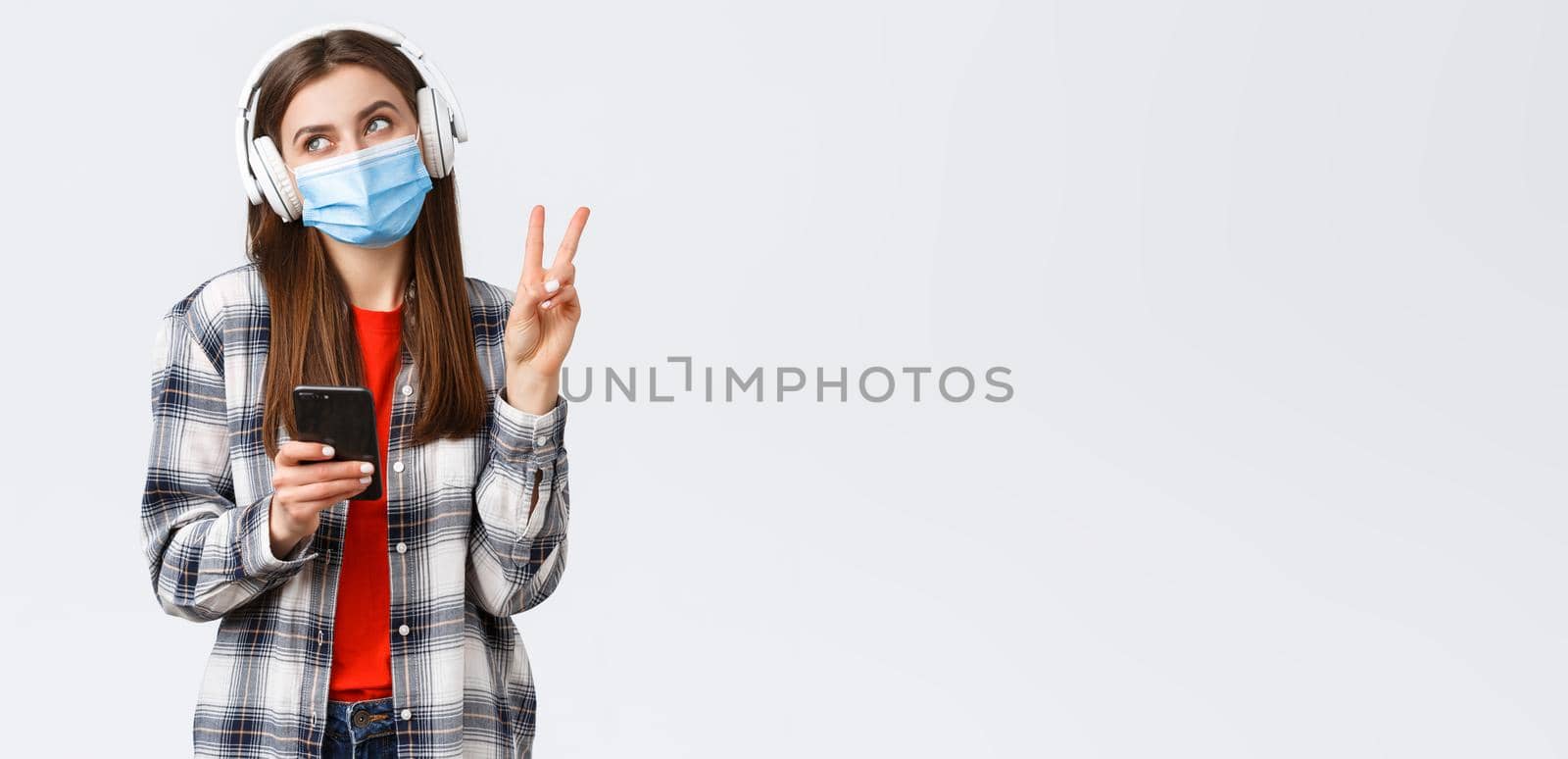 Social distancing, leisure and lifestyle on covid-19 outbreak, coronavirus concept. Happy cheerful silly girl in medical mask and headphones, show peace sign smiling, holding mobile phone by Benzoix