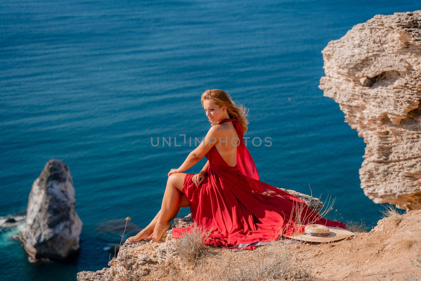 A girl with flowing hair in a long red dress sits on a rock above the sea. The stone can be seen in the sea. by Matiunina