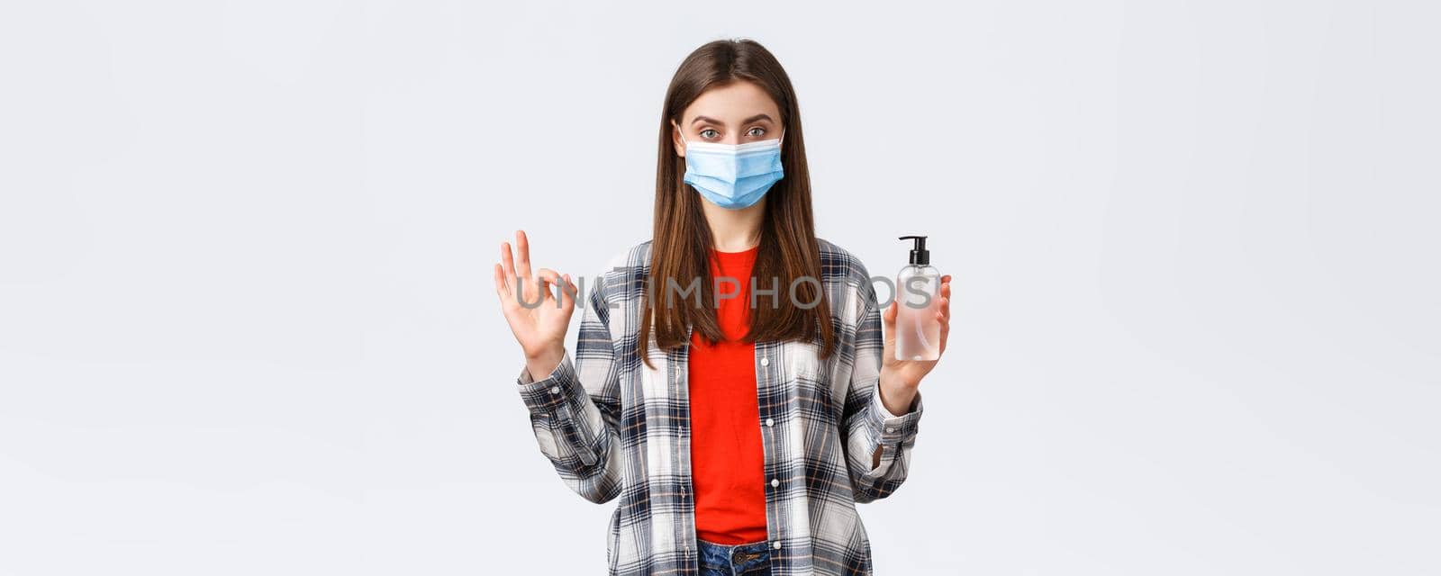 Coronavirus outbreak, leisure on quarantine, social distancing and emotions concept. Pleased assured young woman in medical mask recommend buy this hand sanitizer brand, show okay sign.