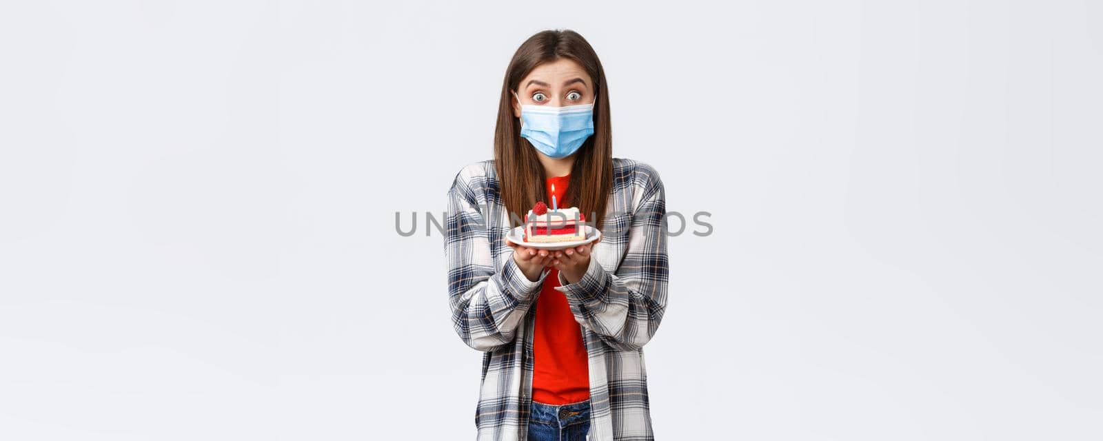 Coronavirus outbreak, lifestyle during social distancing and holidays celebration concept. Cute happy birthday girl making wish, wear medical mask, holding b-day cake, celebrating inside home.