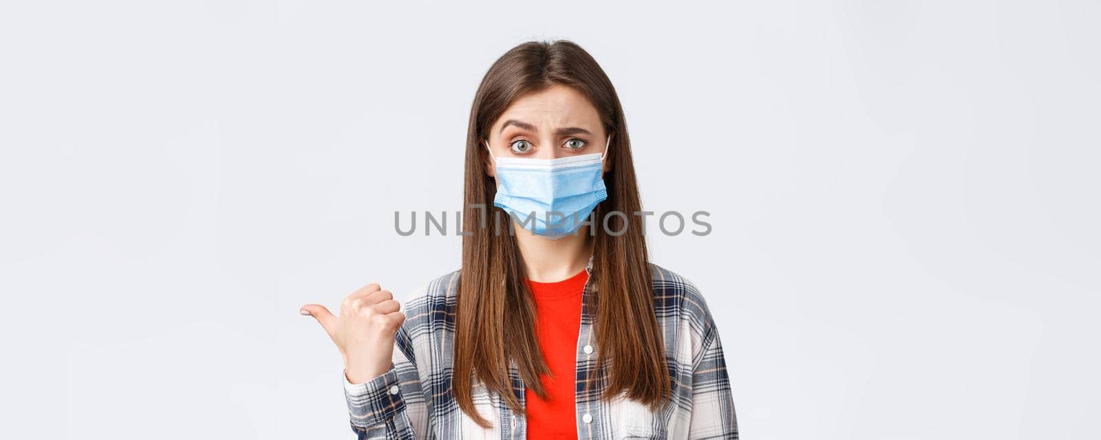 Coronavirus outbreak, leisure on quarantine, social distancing and emotions concept. Skeptical, unimpressed pretty woman in medical mask pointing thumb left and frowning doubtful.
