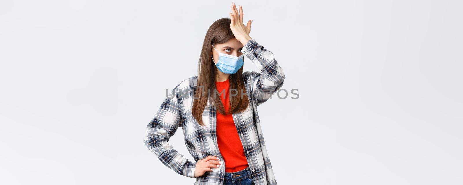 Coronavirus outbreak, leisure on quarantine, social distancing and emotions concept. Bothered and annoyed young woman in medical mask punch forehead and eye roll irritated of smth stupid by Benzoix