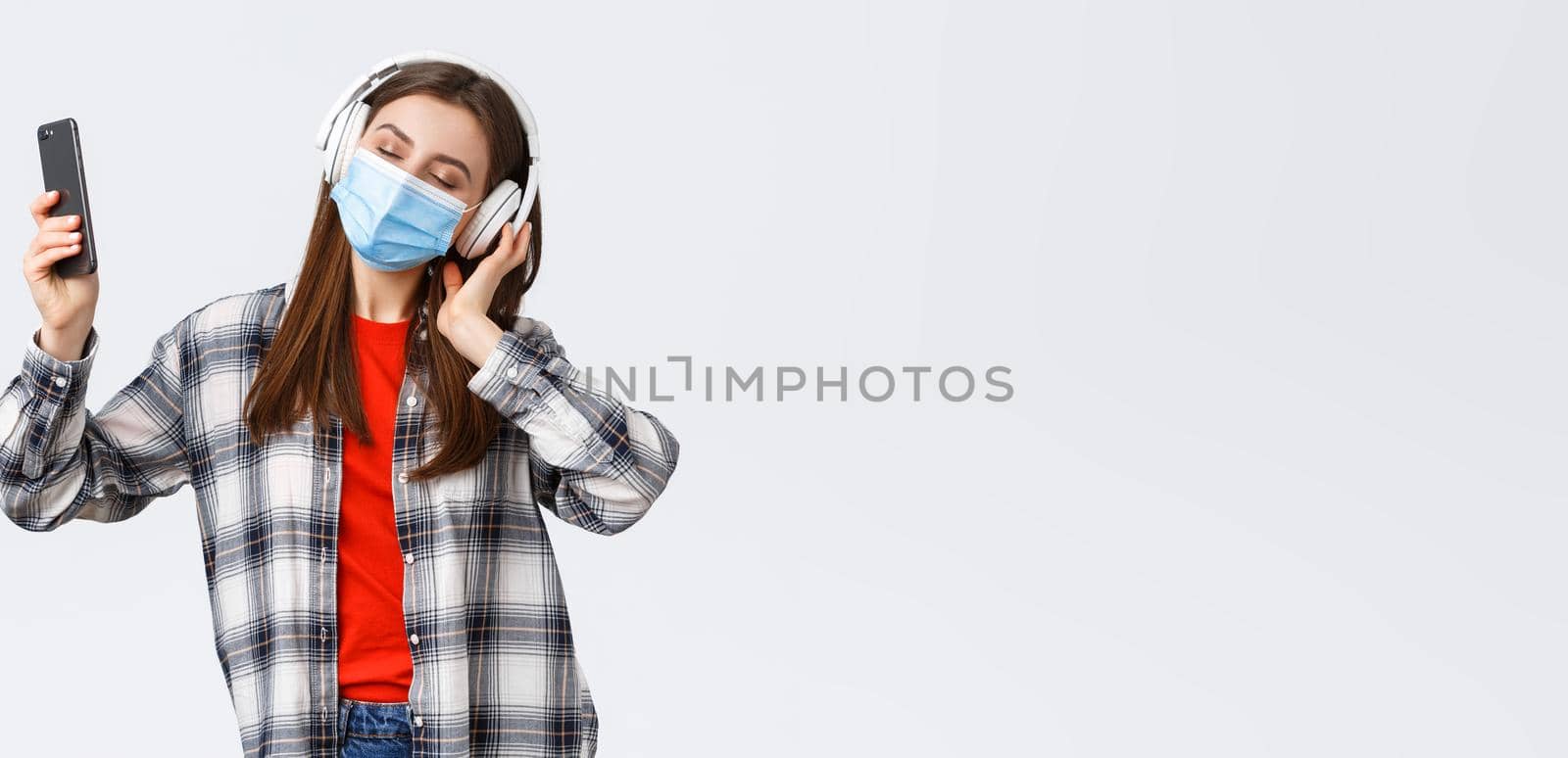 Social distancing, leisure and lifestyle on covid-19 outbreak, coronavirus concept. Carefree young girl in medical mask and headphones, close eyes dancing to music, perfect sound, hold mobile phone.