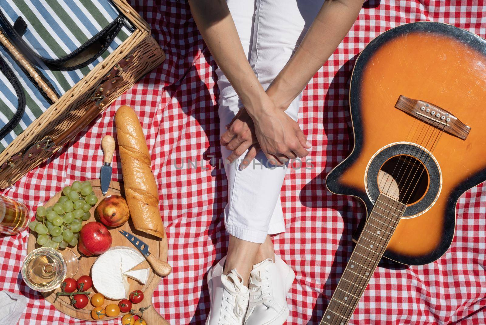 Top view of unrecognizable young woman in white pants outside having picnic, eating and playing guitar. Summer fun and leisure