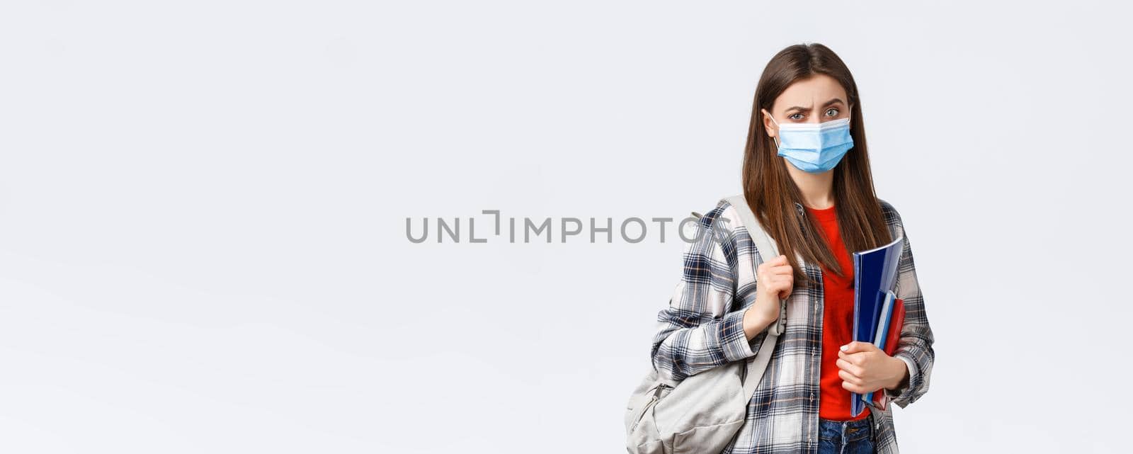 Coronavirus pandemic, covid-19 education, and back to school concept. Doubtful and skeptical female student in medical mask frowning disappointed, hold notebooks and backpack.