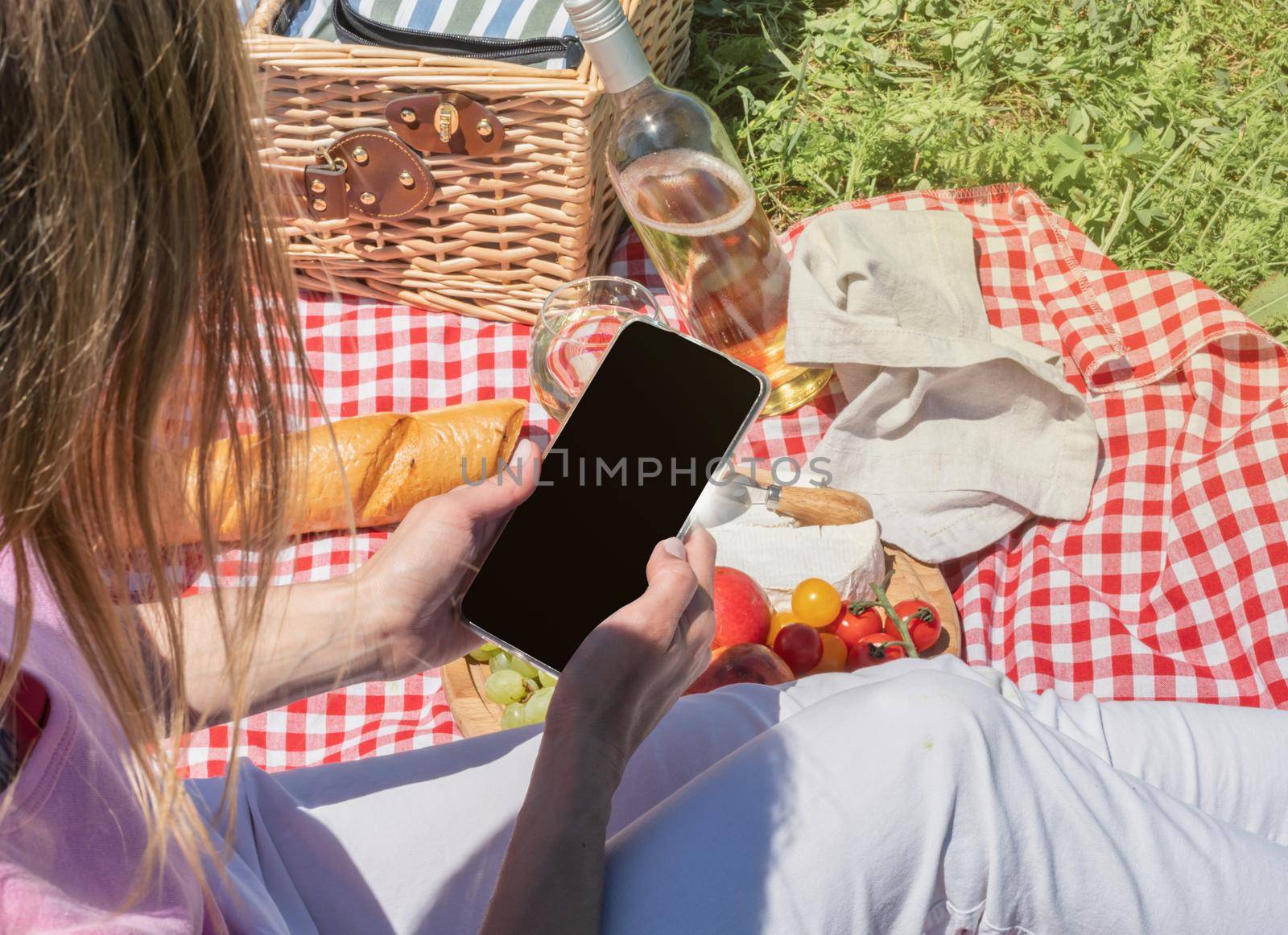 back view of unrecognizable woman in white pants outside having picnic and using smartphone taking photo by Desperada