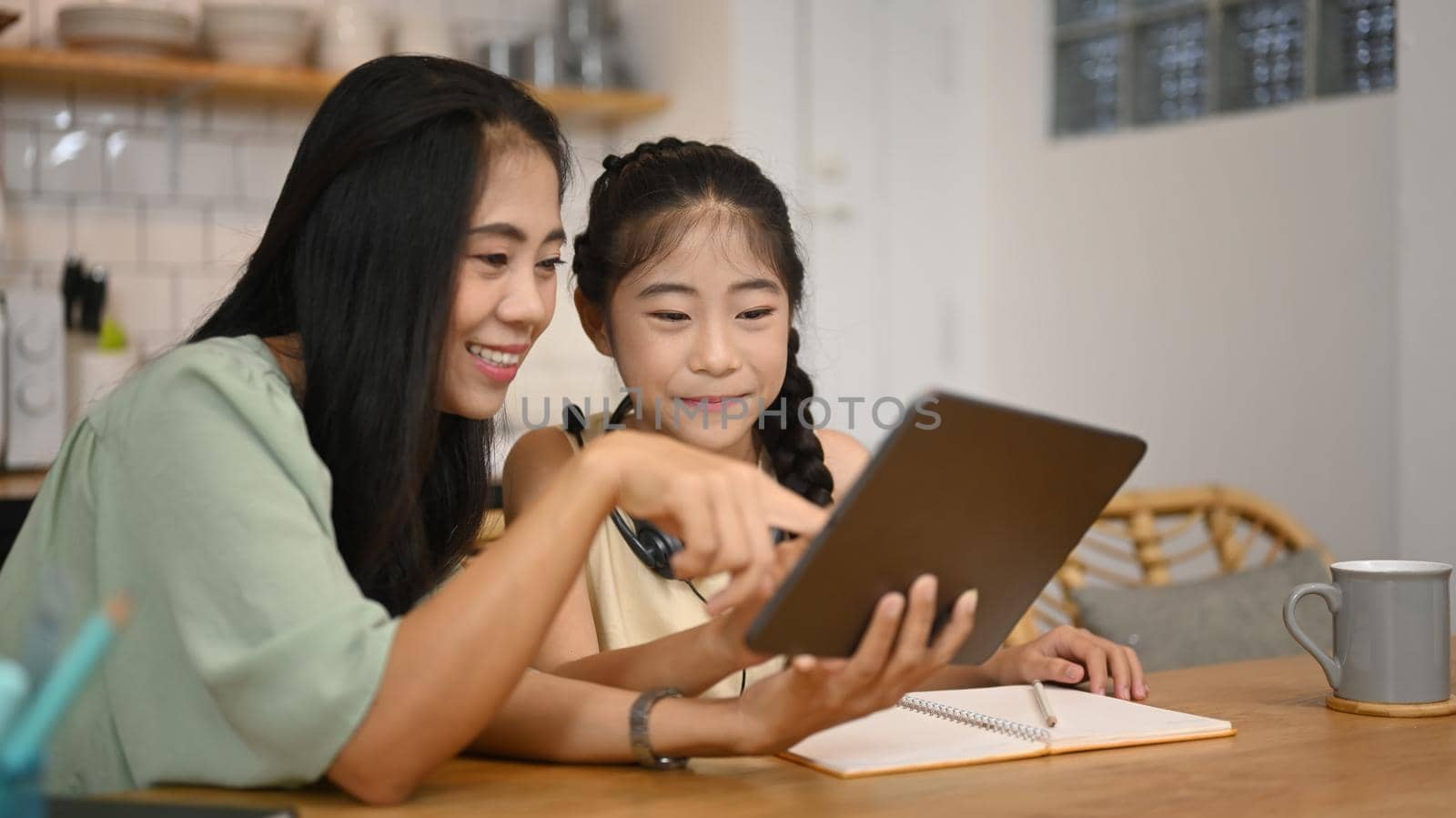 Smiling Asian mother helping her daughter doing homework, surfing internet on digital tablet at home.