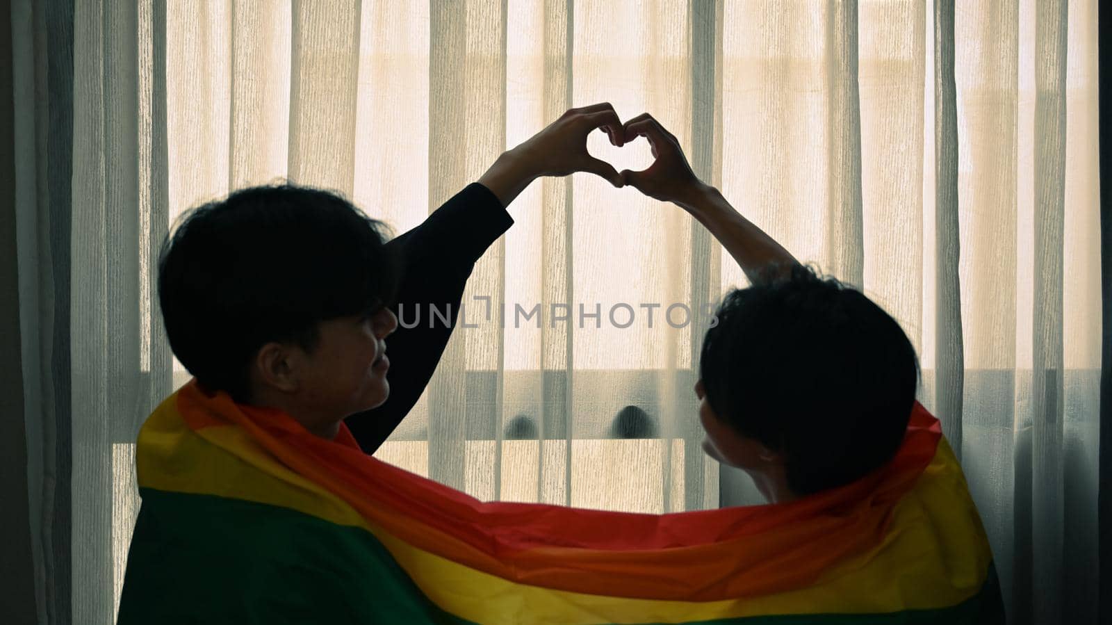 Romantic young gay couple making heart with their hands while sitting under rainbow flag. LGBT, pride, relationships and equality concept.