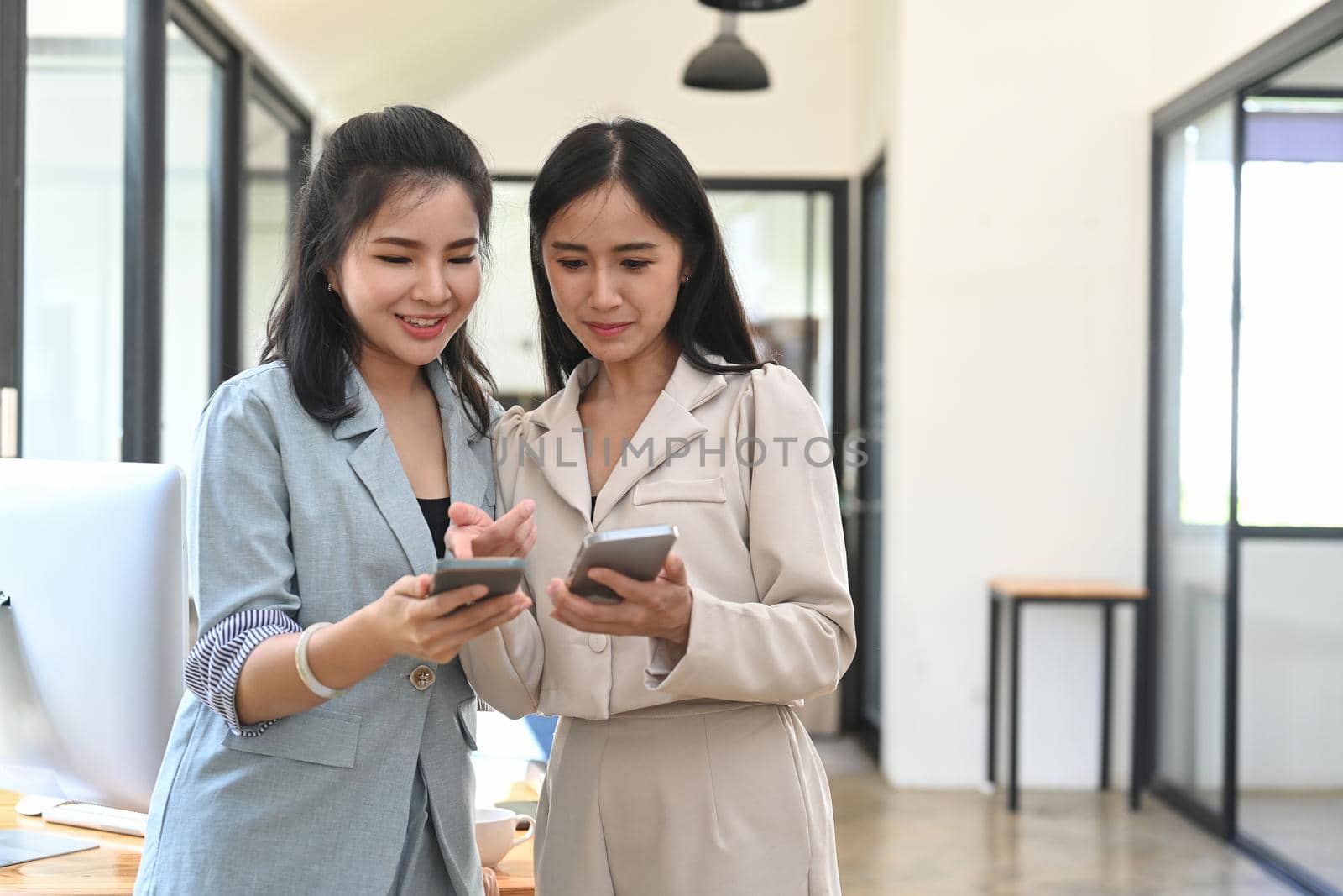 Two Asian business women standing in modern office and looking at smart phone.