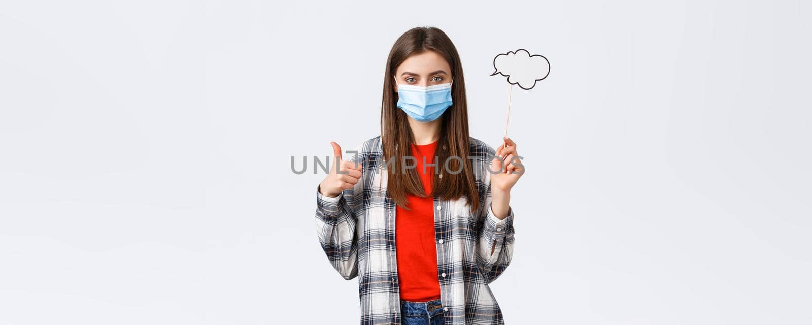 Coronavirus outbreak, leisure on quarantine, social distancing and emotions concept. Pleased young determined girl in medical mask thumb-up, hold bubble commend cloud on stick.